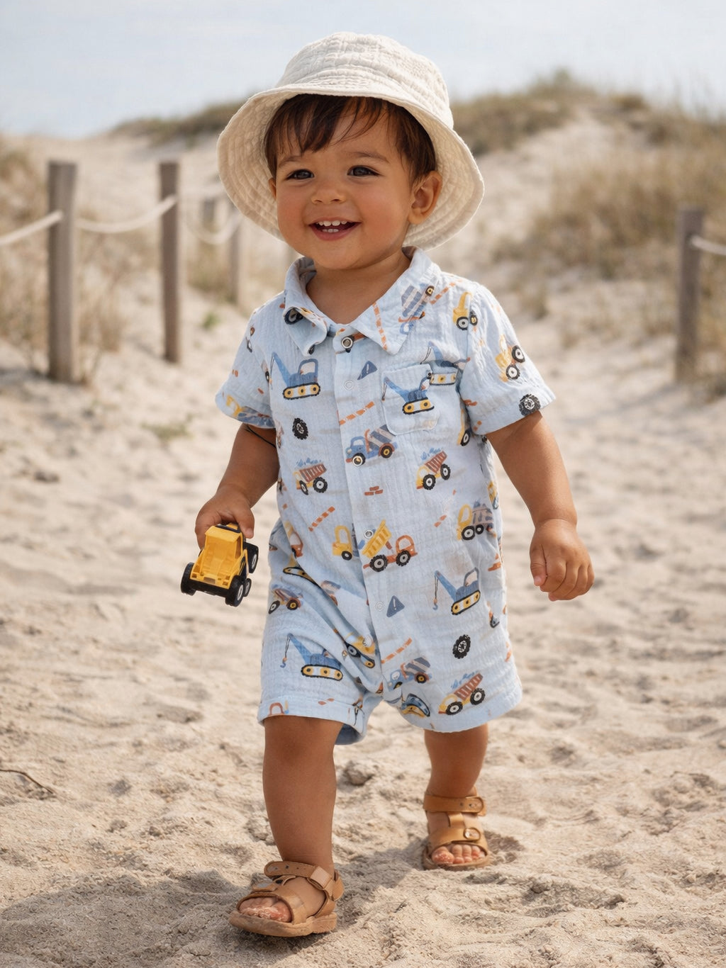 Smiling toddler in a patterned romper, holding a toy truck, walking on a sandy beach with grass in the background.