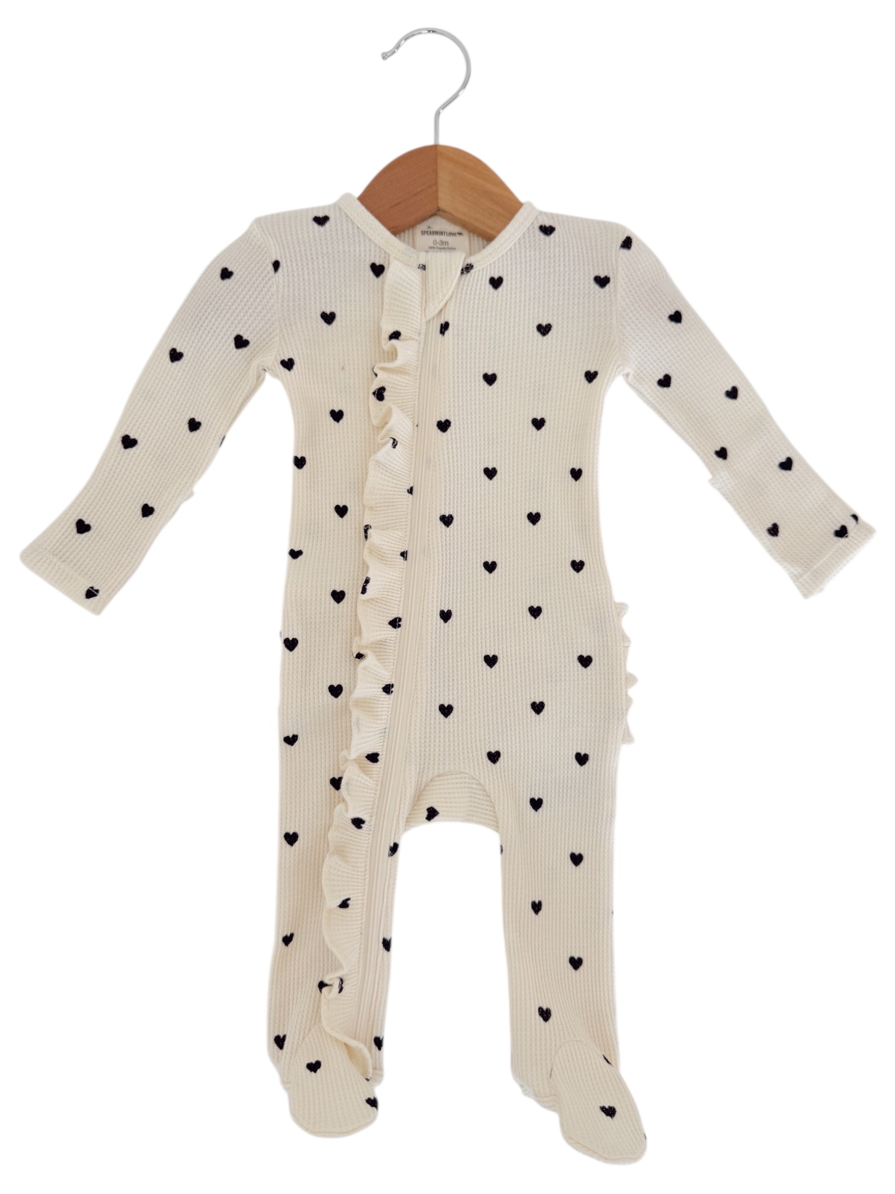 Cream baby onesie with black heart patterns and ruffled front, hanging on a wooden hanger.