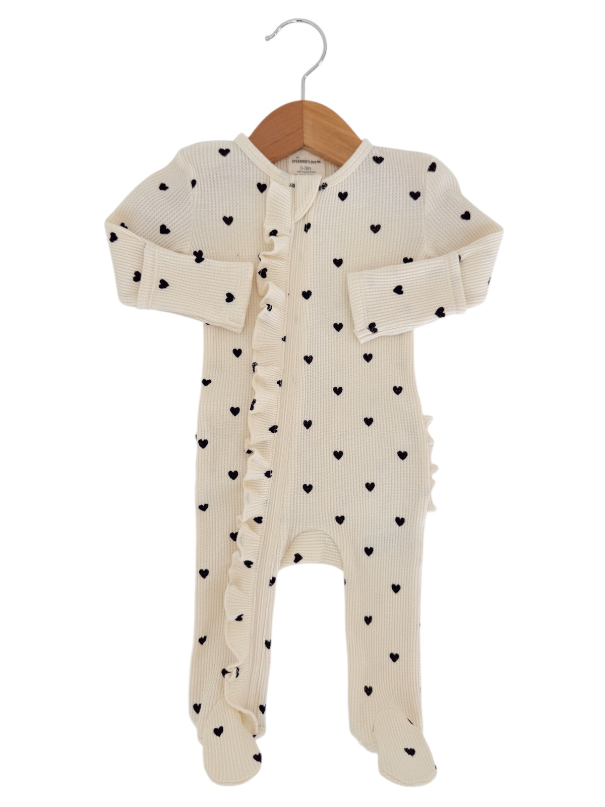 Cream baby onesie with black heart prints and ruffled detailing, hung on a wooden hanger.