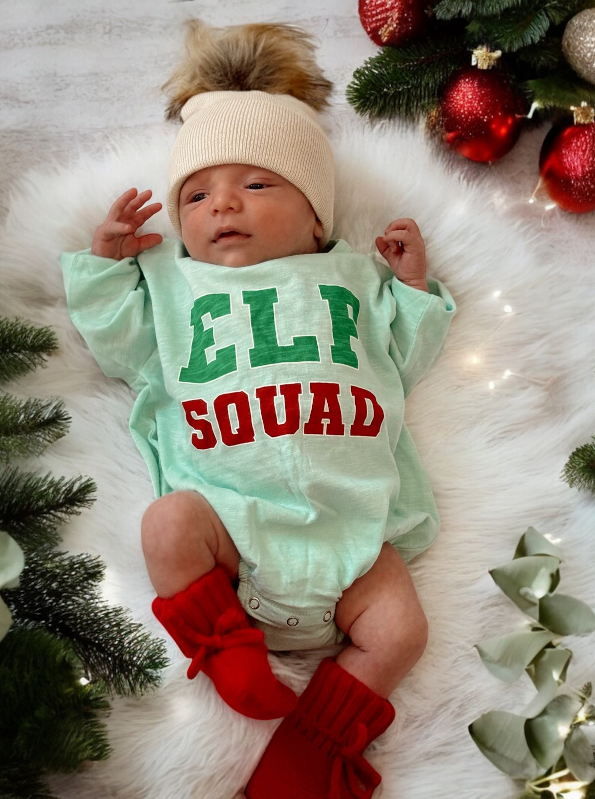 Newborn in an "Elf Squad" outfit with a cream hat and red booties, surrounded by festive greenery and ornaments.