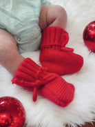Red knitted baby booties on a white fur surface, with festive red ornaments nearby.