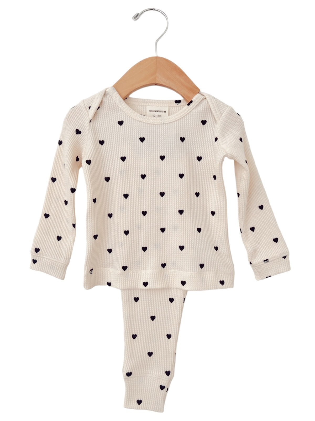 Baby outfit featuring a long-sleeve top and pants with black heart patterns on a white background, hanging on a wooden hanger.