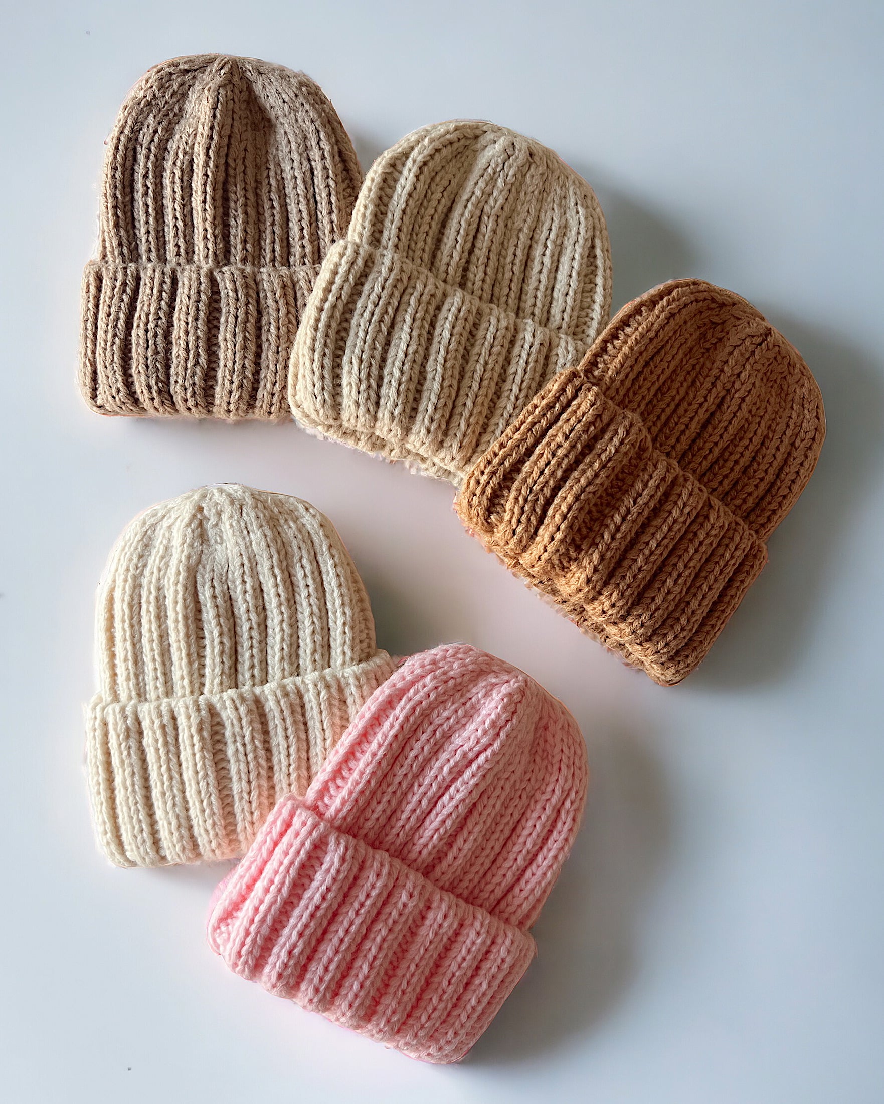 Five knitted beanies in various neutral and pastel colors arranged on a white background.
