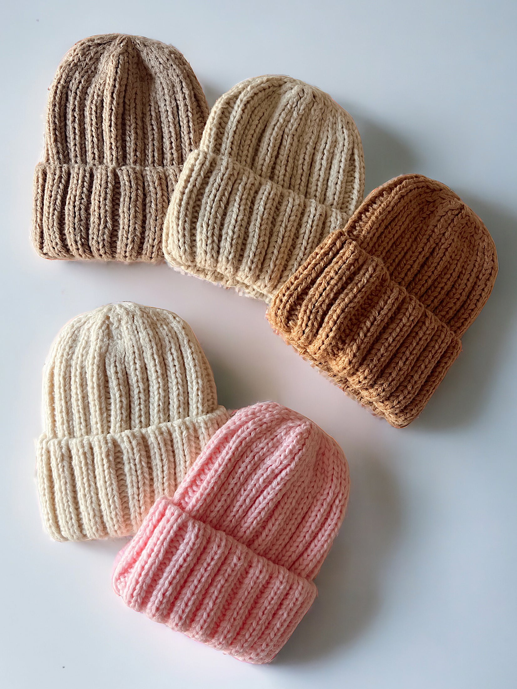 Five knitted beanies in cream, pink, and shades of brown arranged on a white surface.