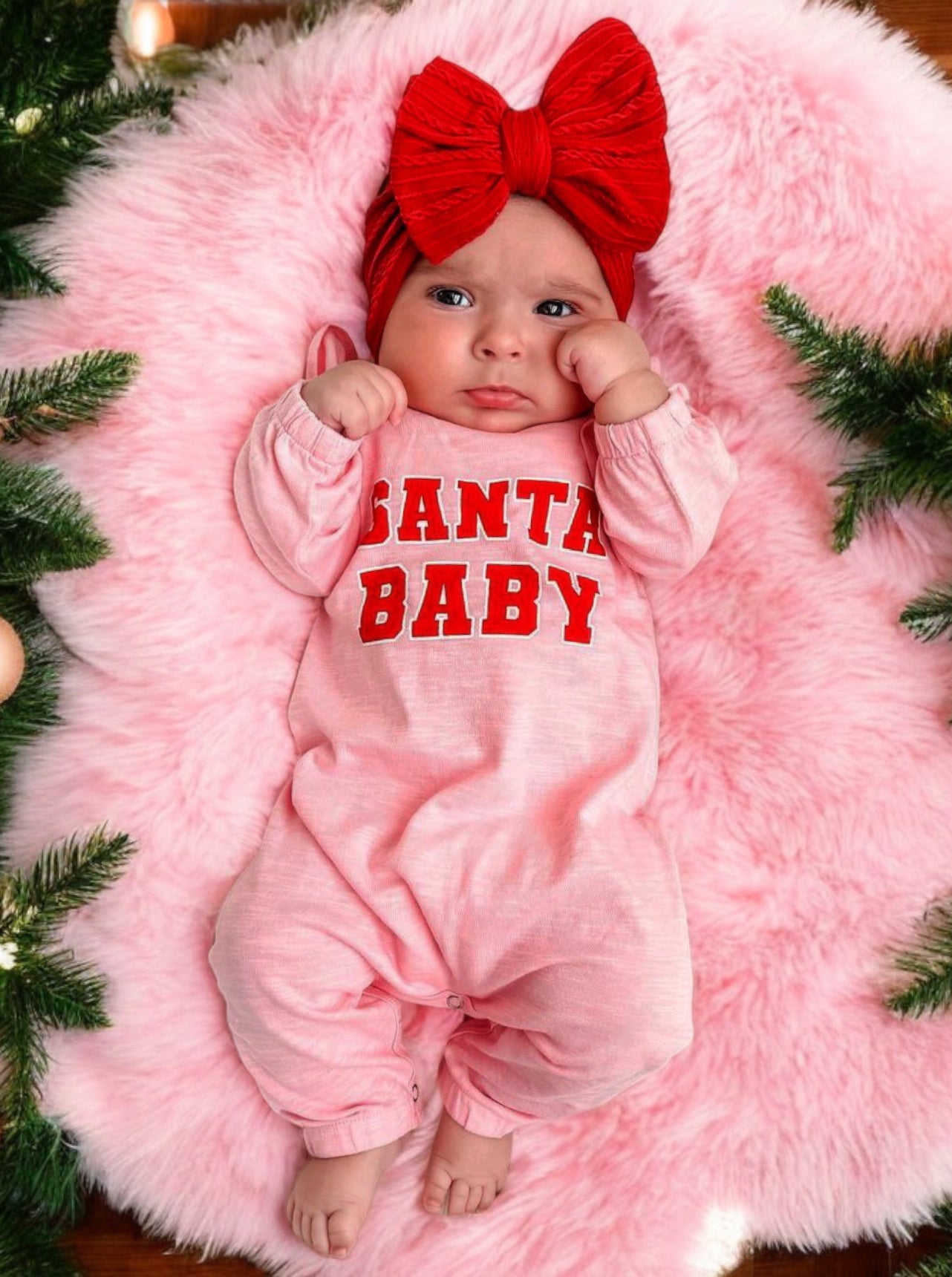 Infant in pink outfit with "Santa Baby" text, wearing a red bow, on a fluffy pink rug surrounded by Christmas greenery.