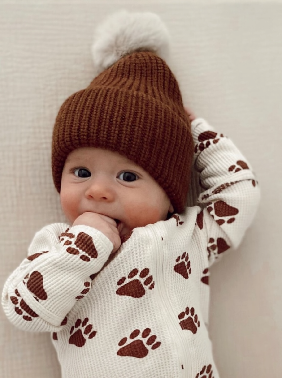 Baby wearing a brown knit hat and paw-print sweater, smiling and sucking on their hand.