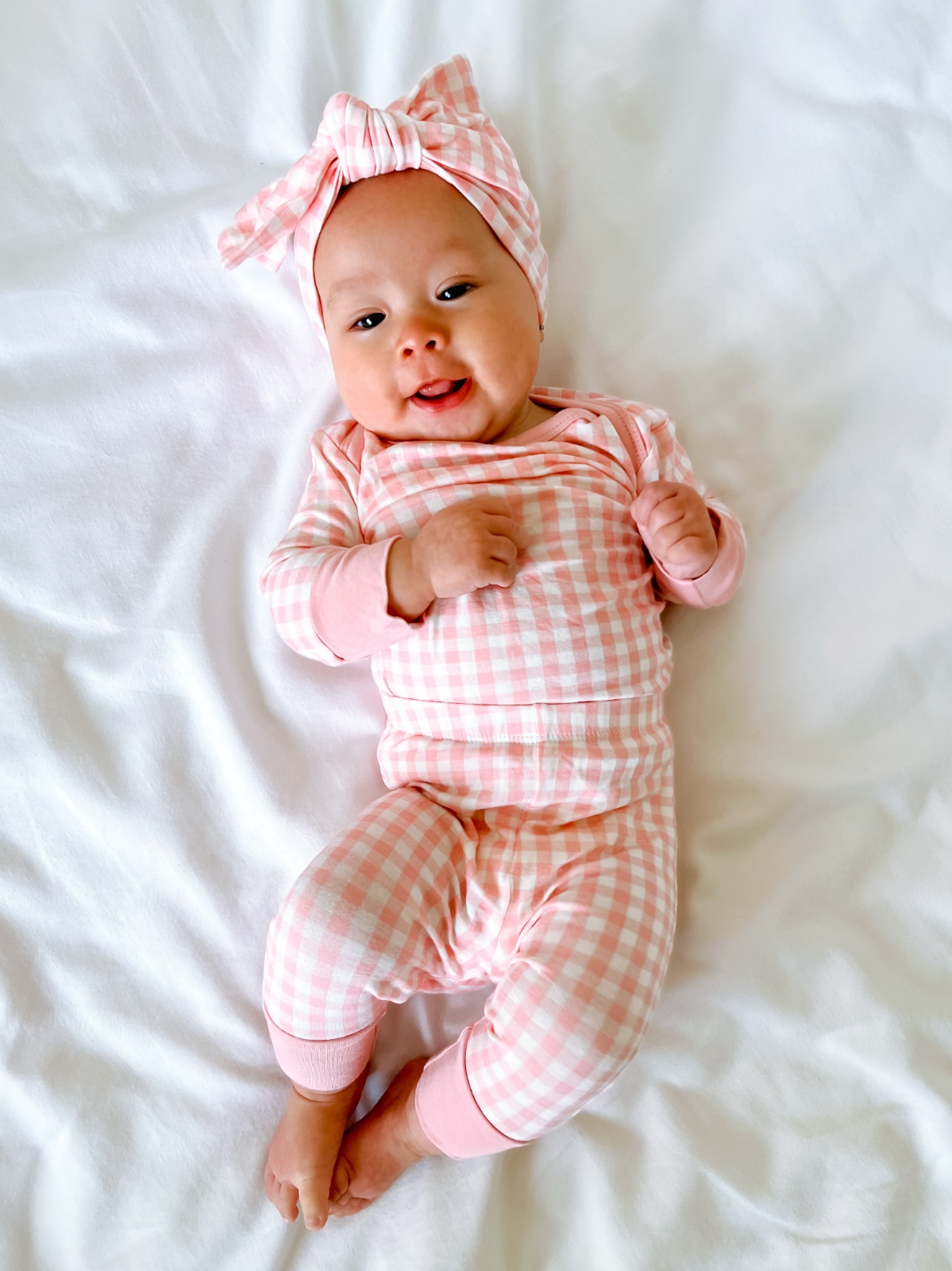 Smiling baby in a pink checkered outfit and matching headband, lying on a white blanket.