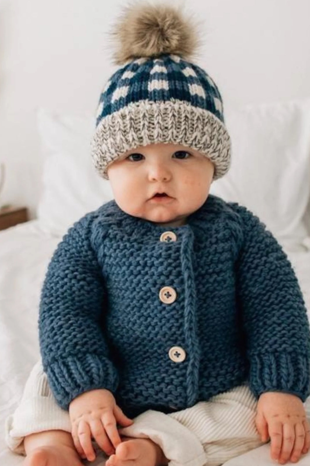 Baby in a cozy blue knitted sweater and a plaid hat with a pom-pom, sitting on a bed with white bedding.