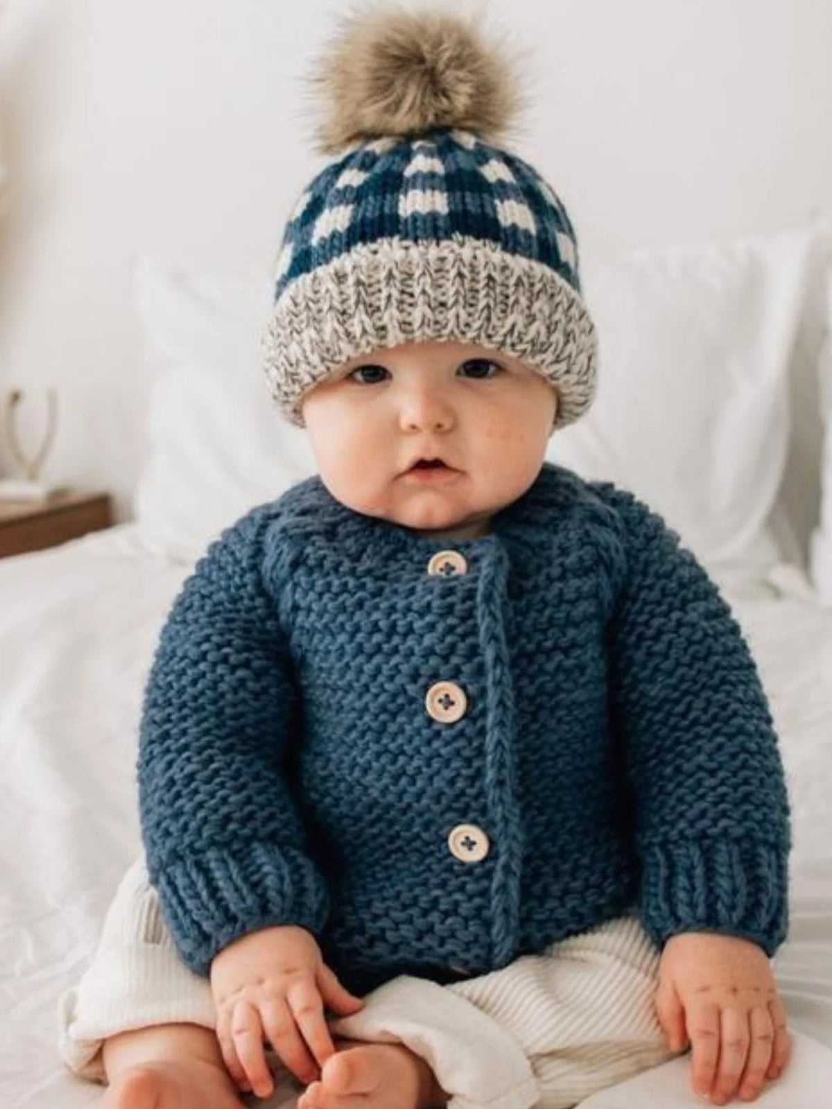 Baby in a cozy blue knitted sweater and a plaid hat with a pom-pom, sitting on a bed with white bedding.