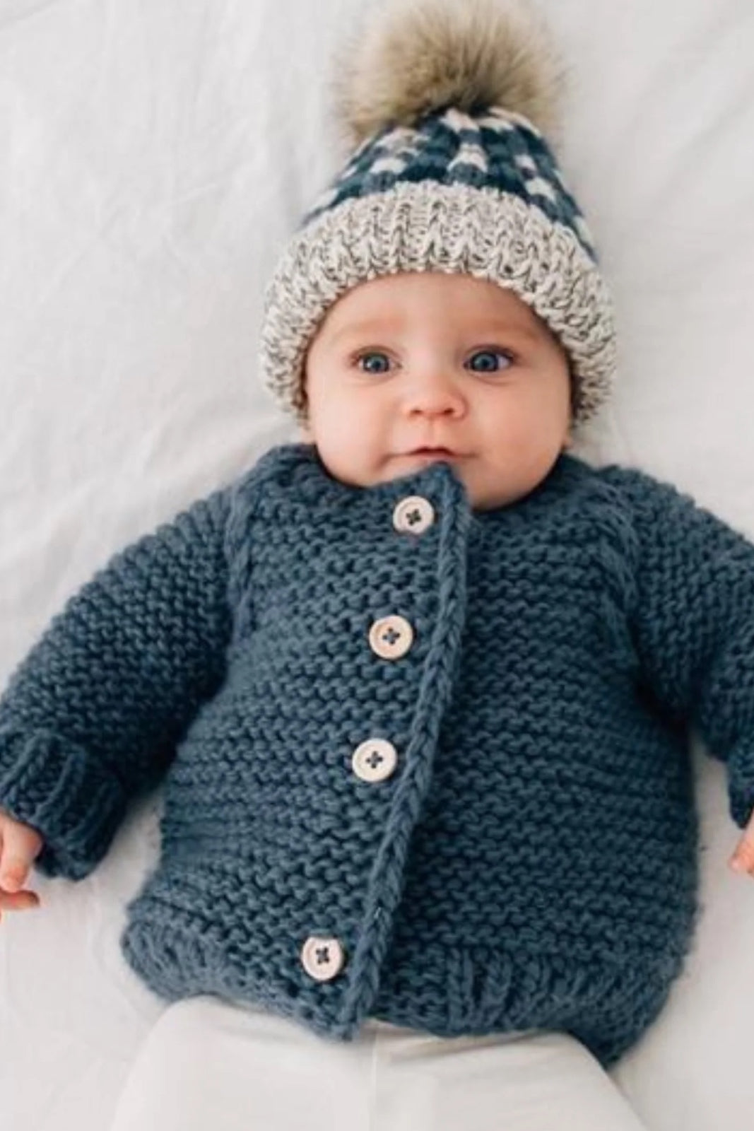 Baby wearing a cozy knitted gray jacket and a patterned hat with a pom-pom, resting on a white background.