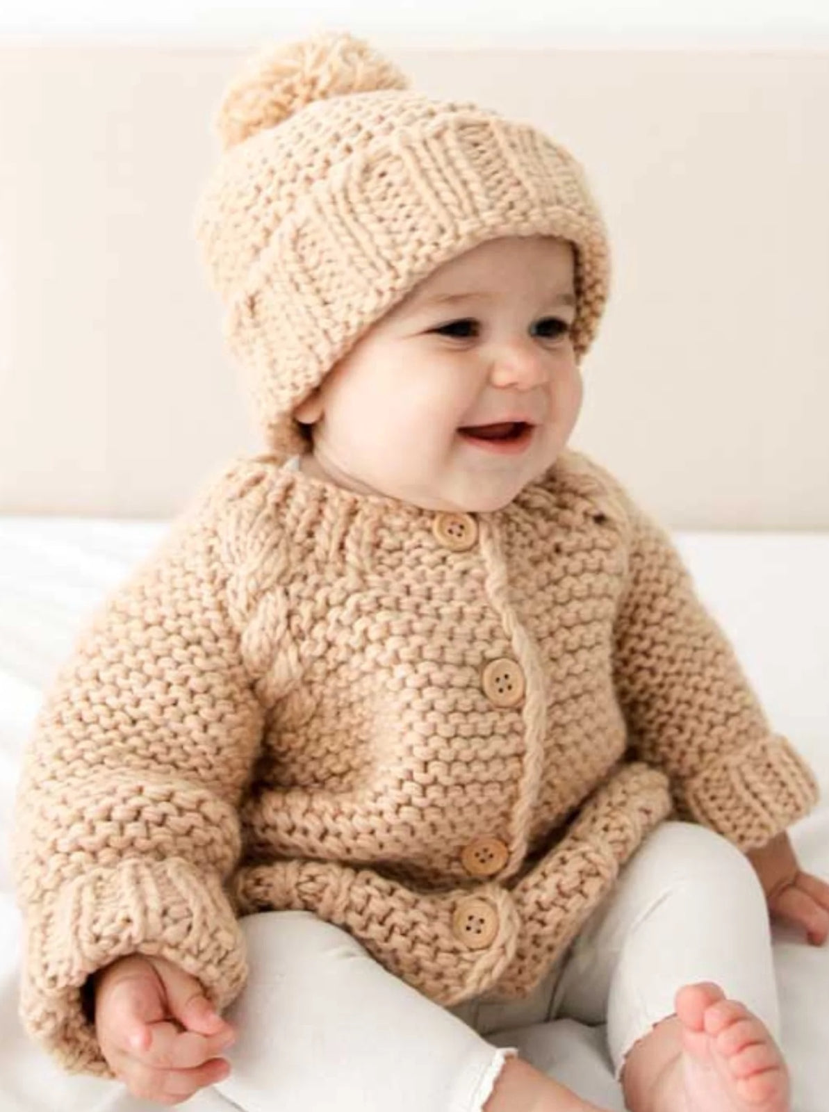 Smiling baby wearing a knitted beige sweater and hat, sitting on a light-colored surface.