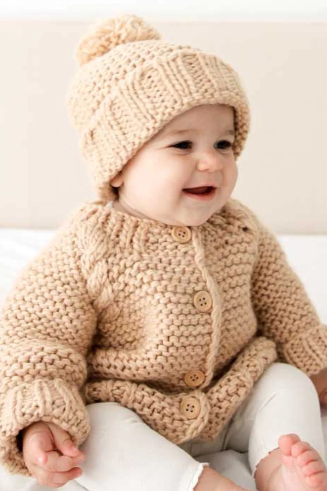 Smiling baby wearing a knitted beige sweater and hat, sitting on a light-colored surface.