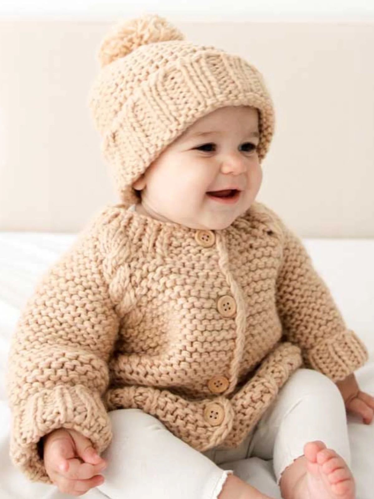 Smiling baby wearing a knitted beige sweater and hat, sitting on a light-colored surface.