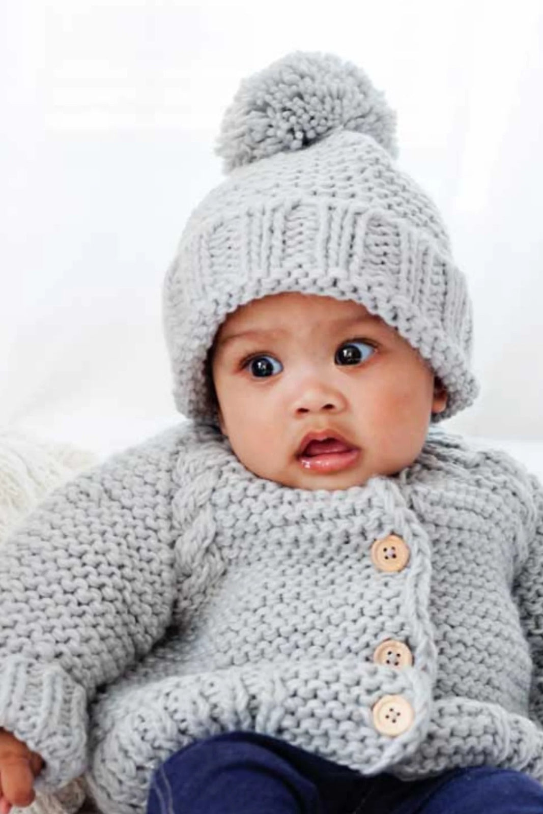 Baby wearing a cozy gray knitted sweater and pom-pom hat, looking curiously at the camera.