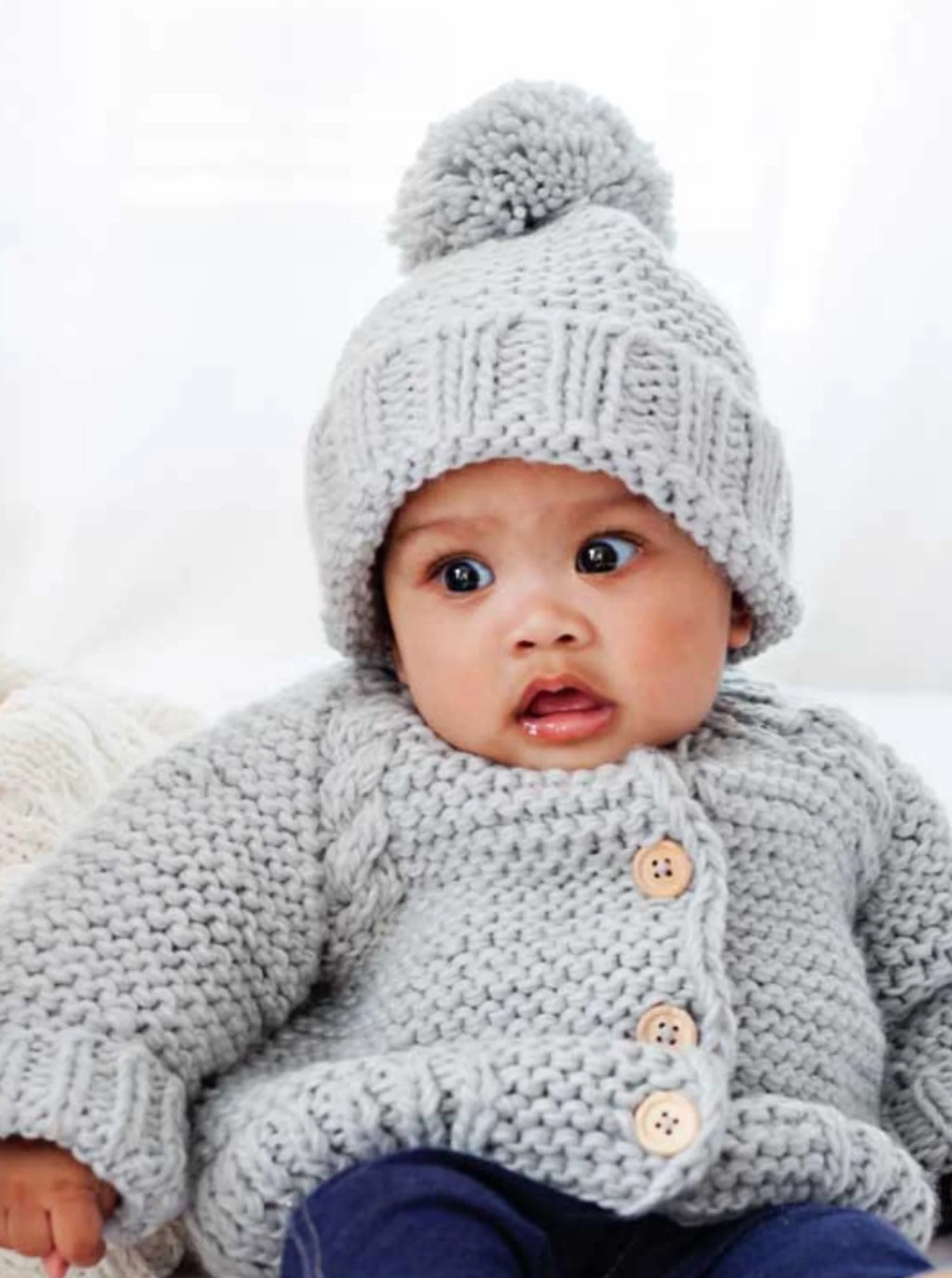 Baby wearing a cozy gray knitted sweater and hat, with a surprised expression, sitting on a soft surface.