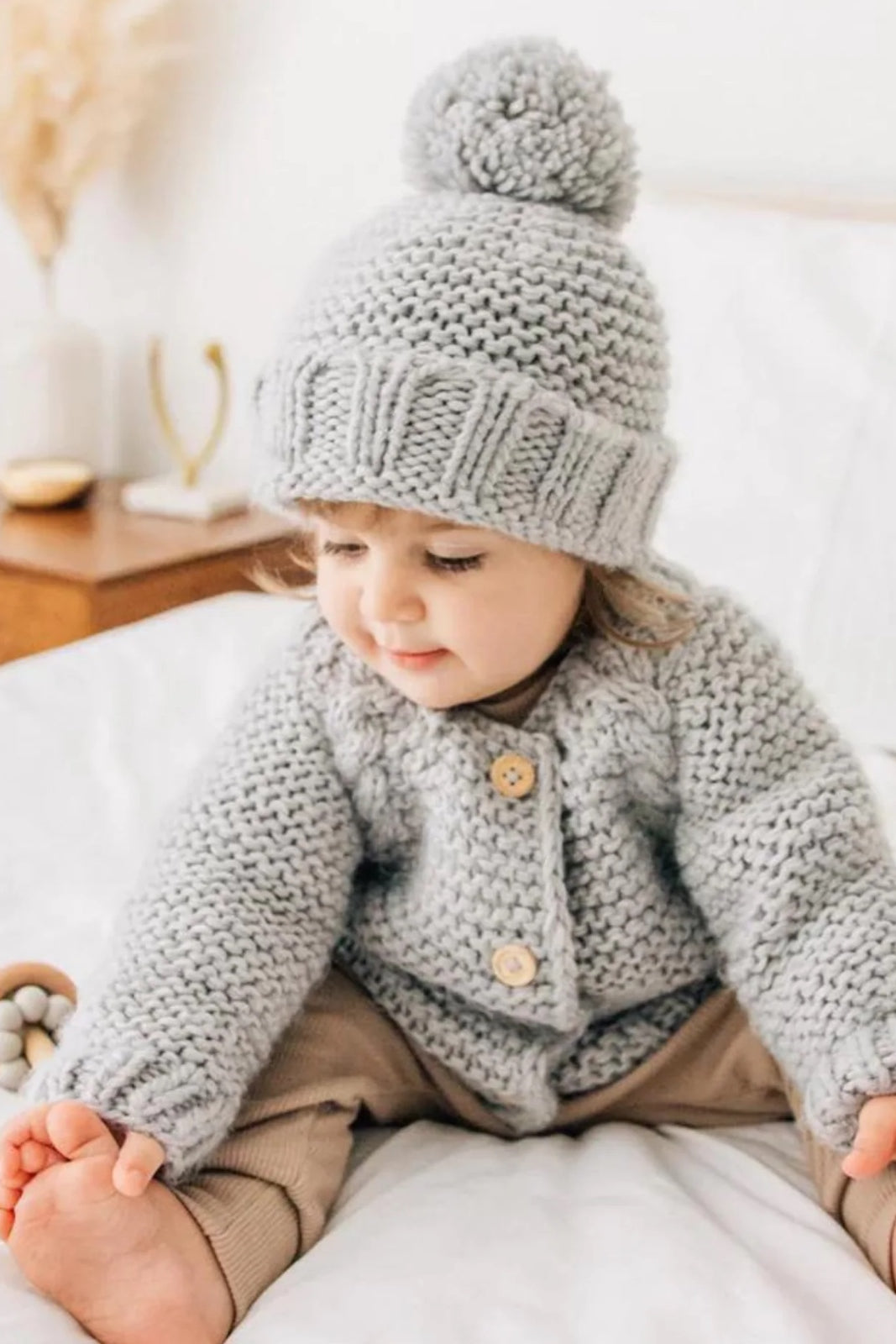 Toddler in a cozy gray knitted cardigan and hat, sitting on a bed with a playful expression.