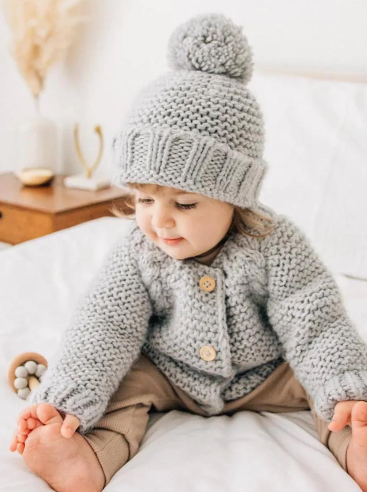 Baby in a cozy gray knit sweater and hat, sitting on a bed with a playful expression. Soft, neutral decor in background.