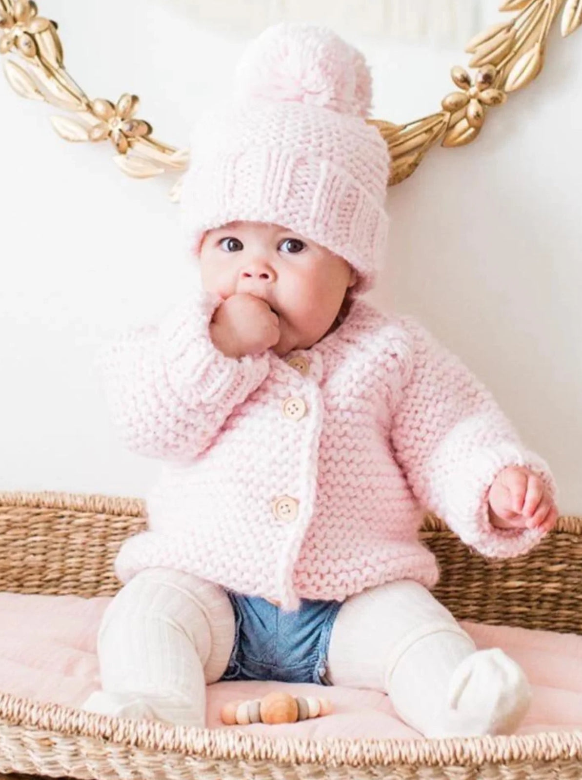 Baby in a pink knitted sweater and hat sits on a woven basket, sucking thumb, with pastel toys nearby.