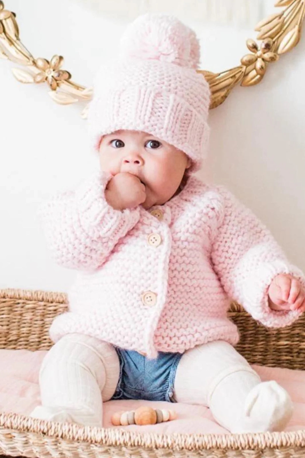 Baby in a pink knitted sweater and hat sits on a woven basket, sucking thumb, with pastel toys nearby.