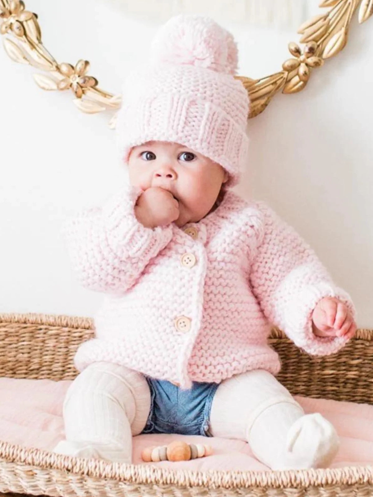 Baby in a pink knitted sweater and hat sits on a woven basket, sucking thumb, with pastel toys nearby.
