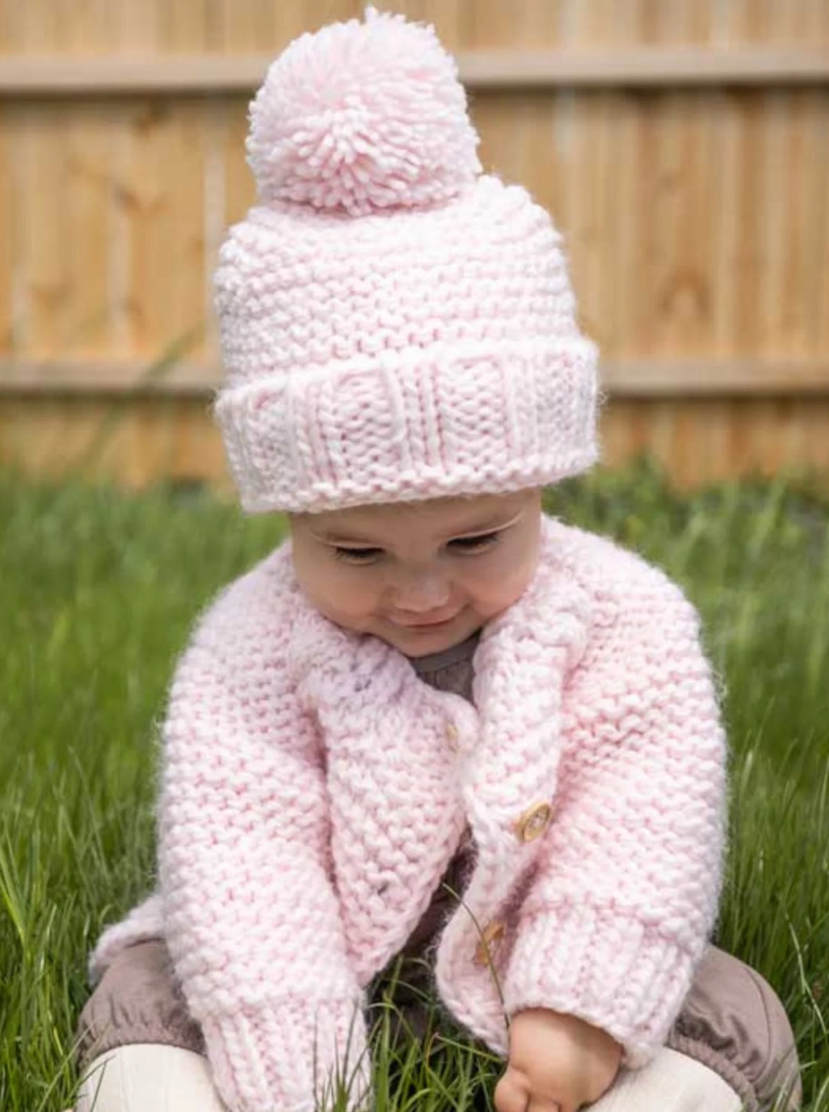 Baby sitting in grass, wearing a pink knitted hat with pom-pom and a matching cardigan, looking down.
