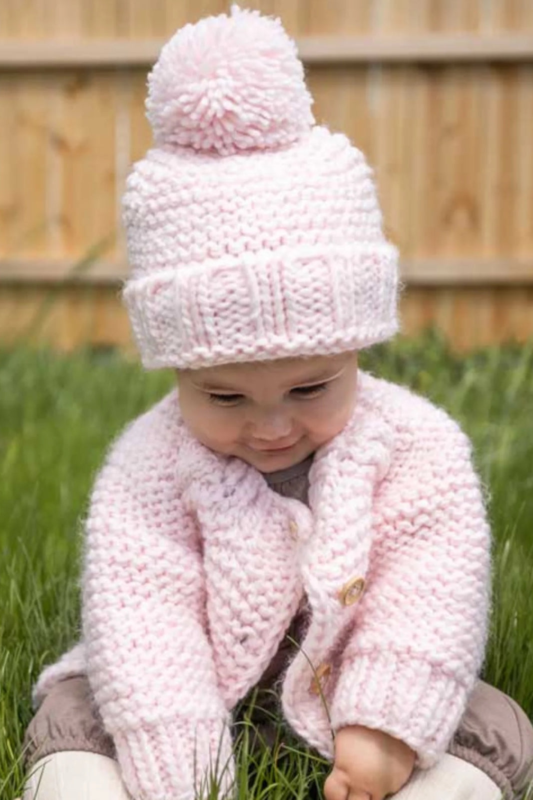 Baby sitting in grass, wearing a pink knitted hat with pom-pom and a matching cardigan, looking down.