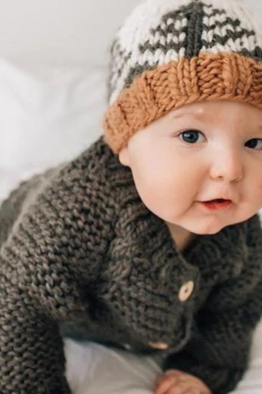 Baby wearing a knitted sweater and hat, smiling while crawling on a bed with white sheets.