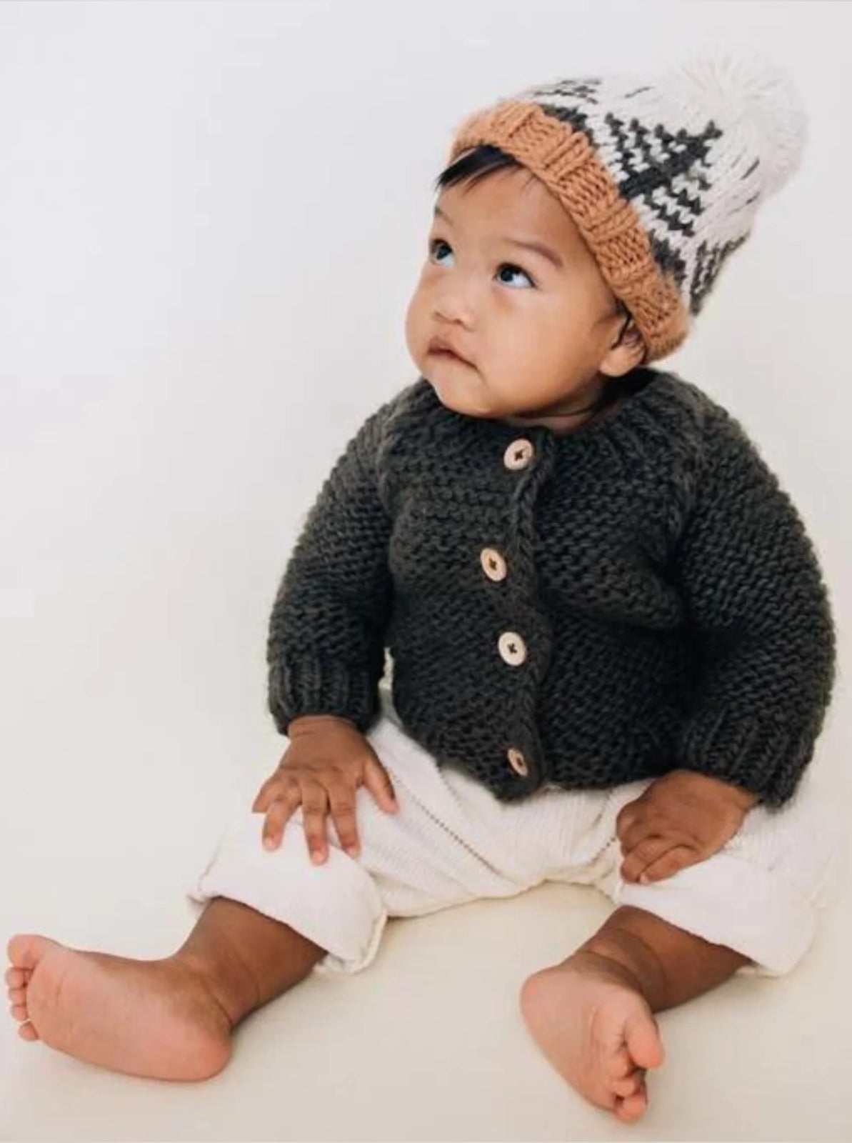 Baby sitting on the floor, wearing a cozy knitted cardigan and a hat, looking up with a curious expression.