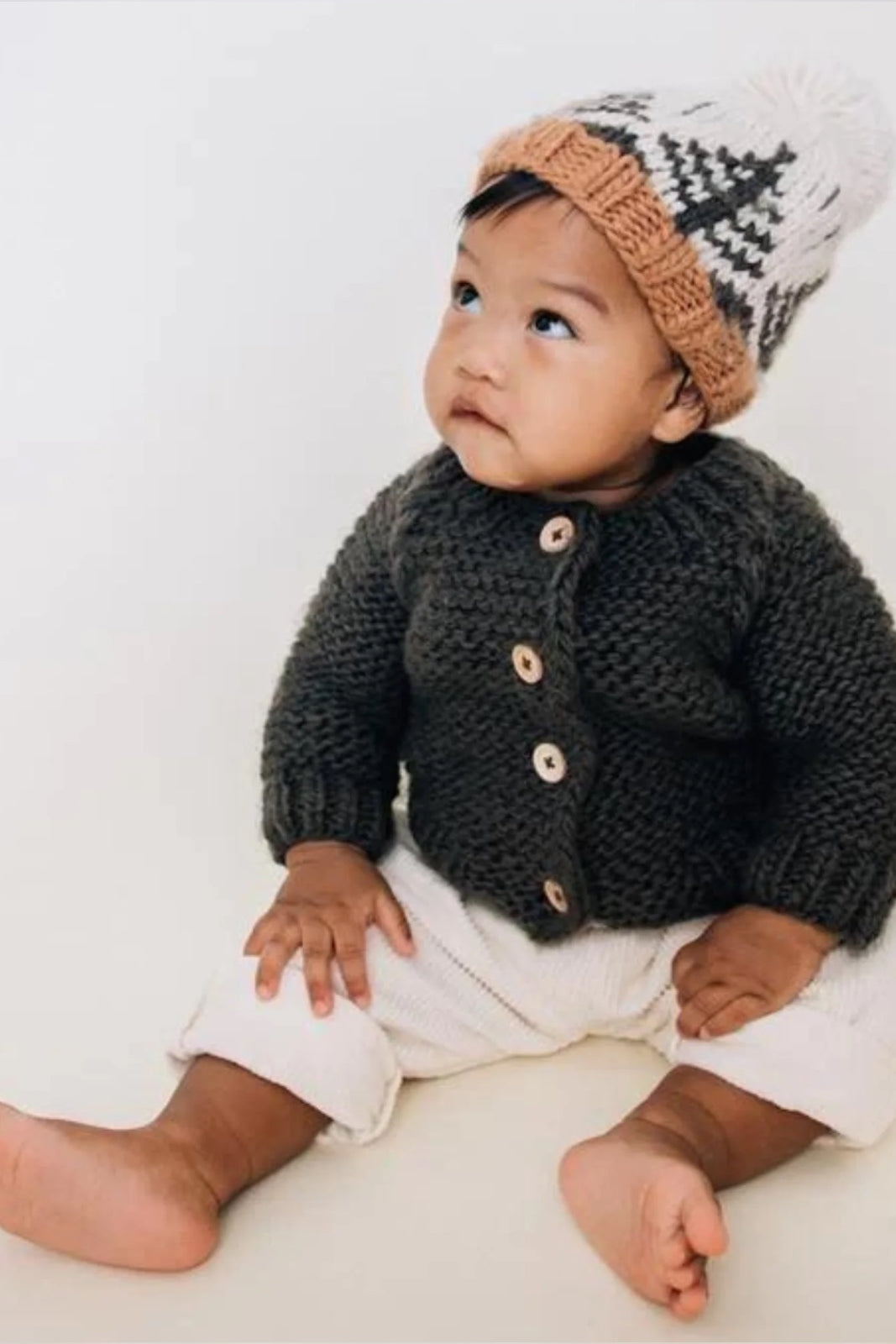 Baby sitting on the floor, wearing a cozy knitted cardigan and a hat, looking up with a curious expression.