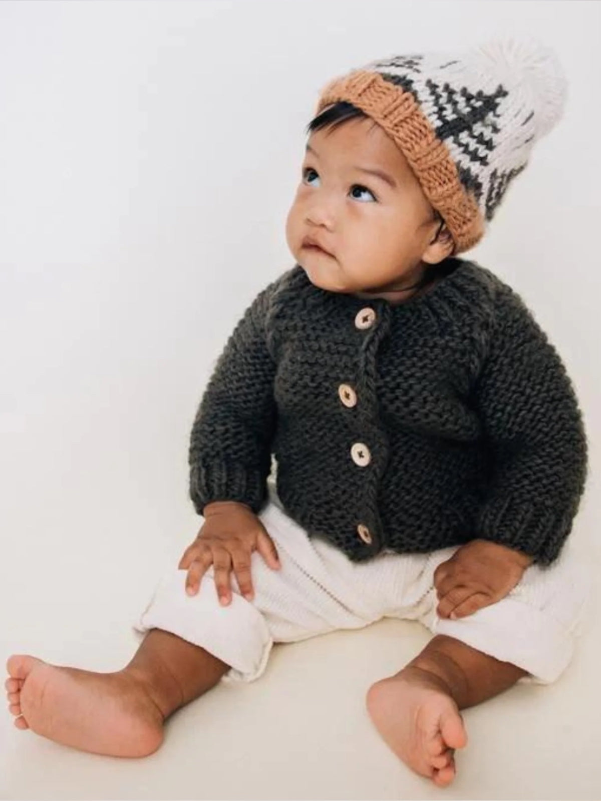 Baby sitting on the floor, wearing a cozy knitted cardigan and a hat, looking up with a curious expression.