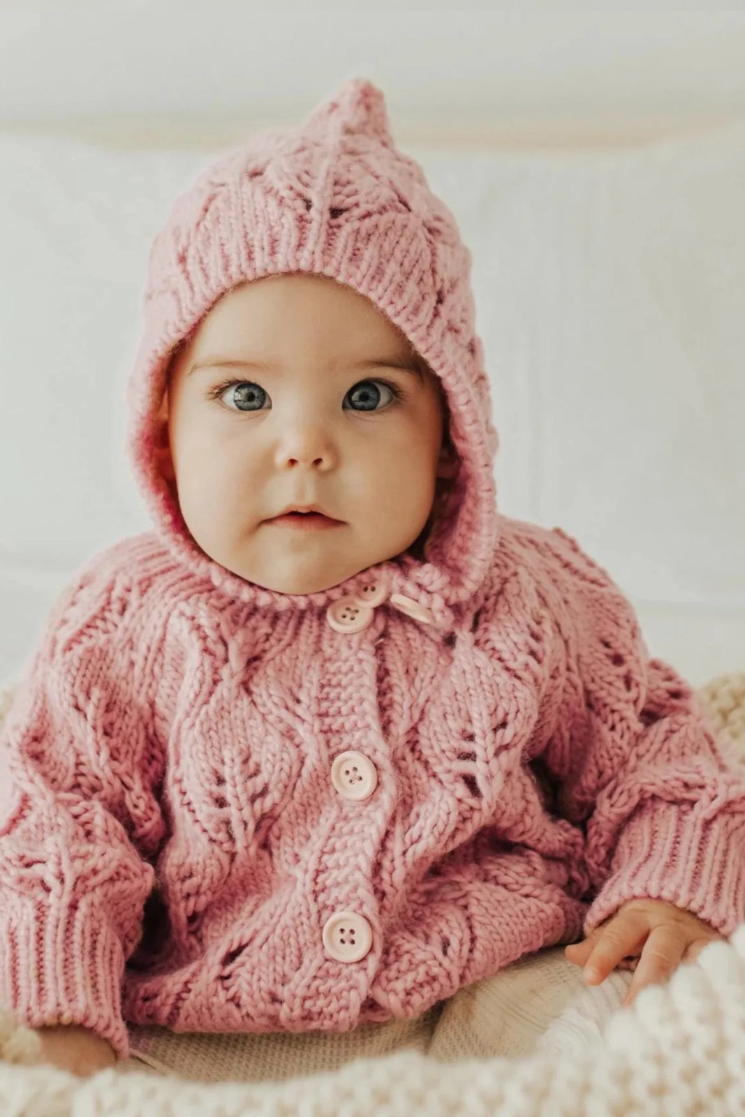 Baby in a pink knitted sweater and bonnet, sitting on a cozy blanket with a neutral background.