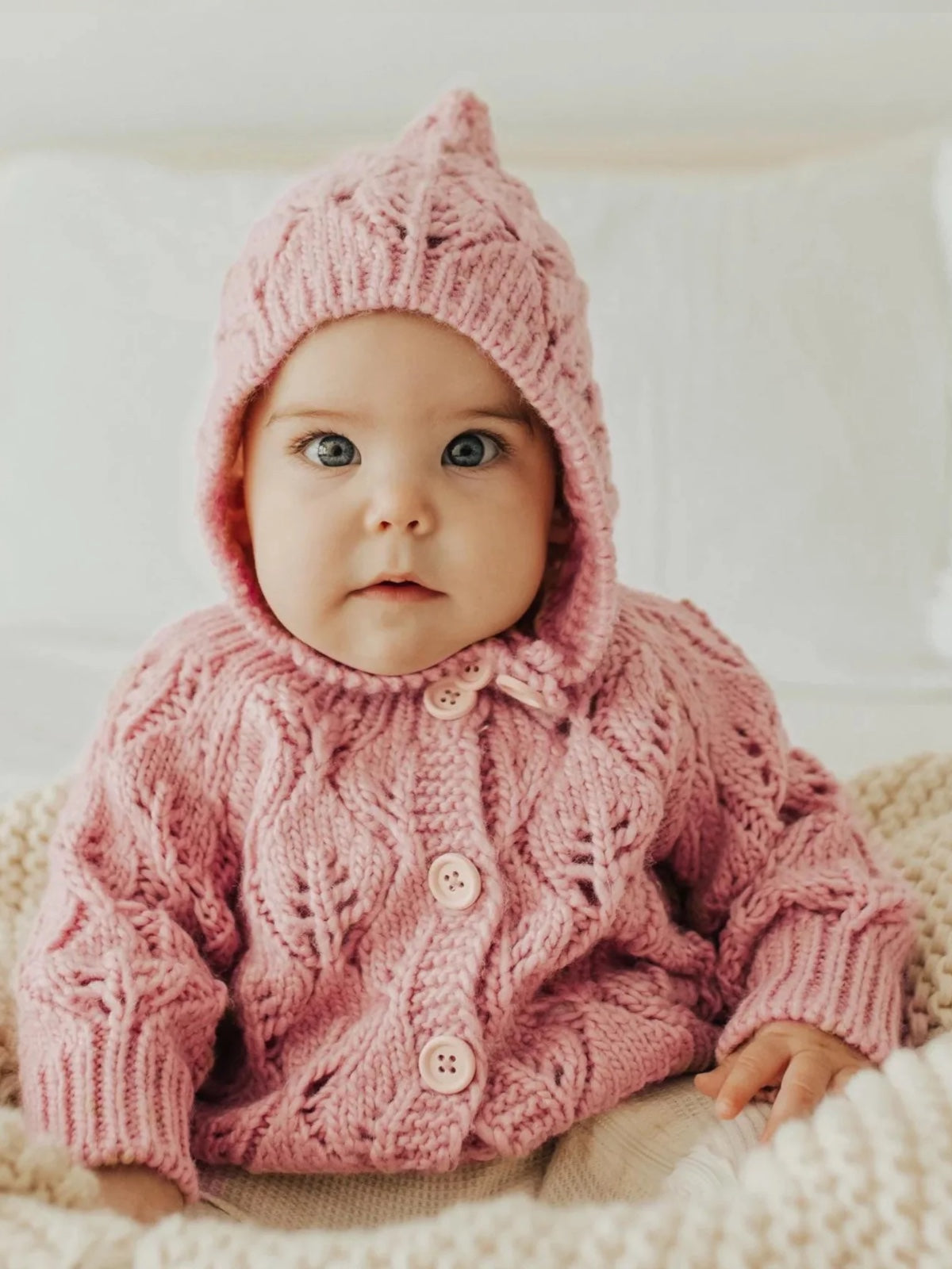 Baby in a pink knitted sweater and bonnet, sitting on a cozy blanket with a neutral background.