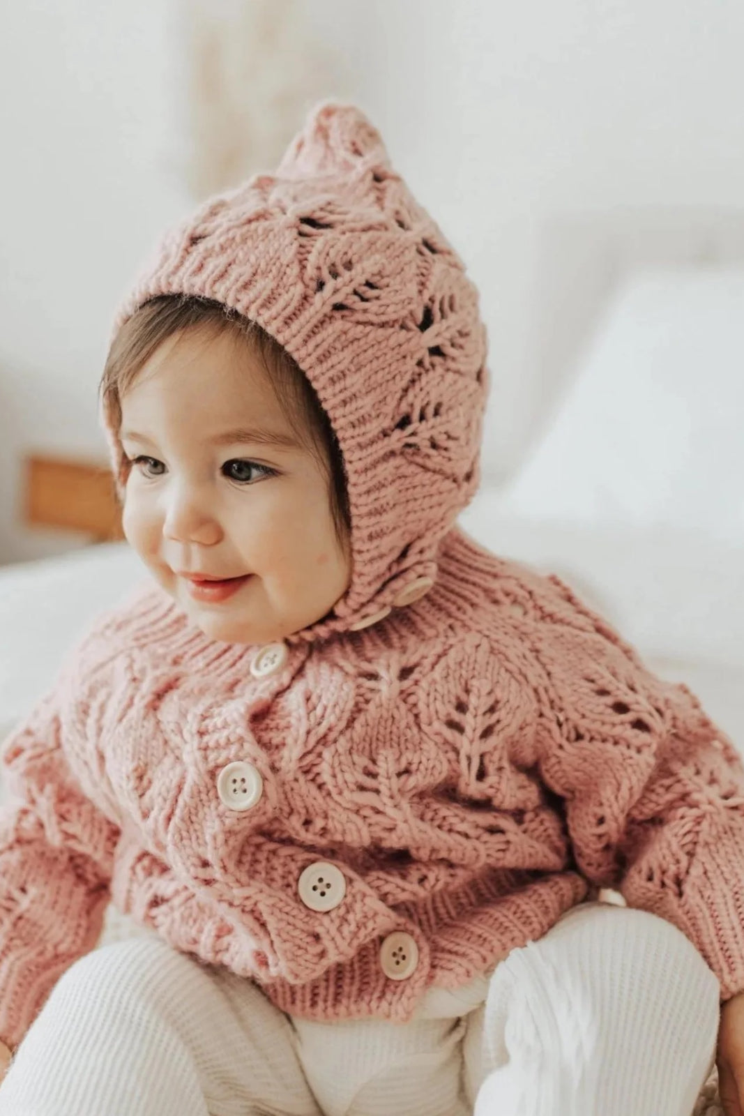 Smiling toddler in a pink knitted sweater and matching hat, sitting on a bed with a cozy backdrop.