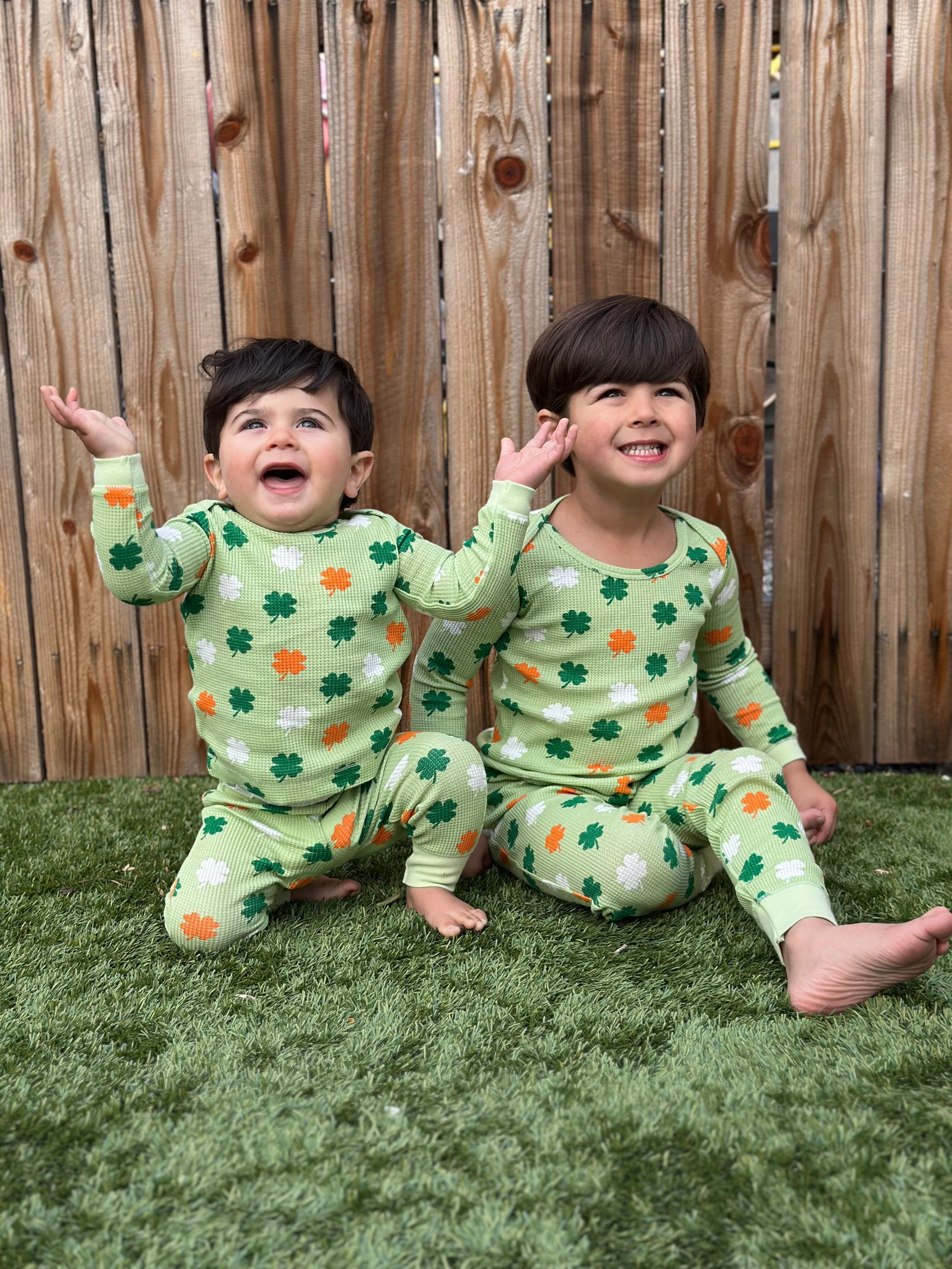 Two children in matching green pajama sets with shamrock patterns, sitting on grass and smiling joyfully.