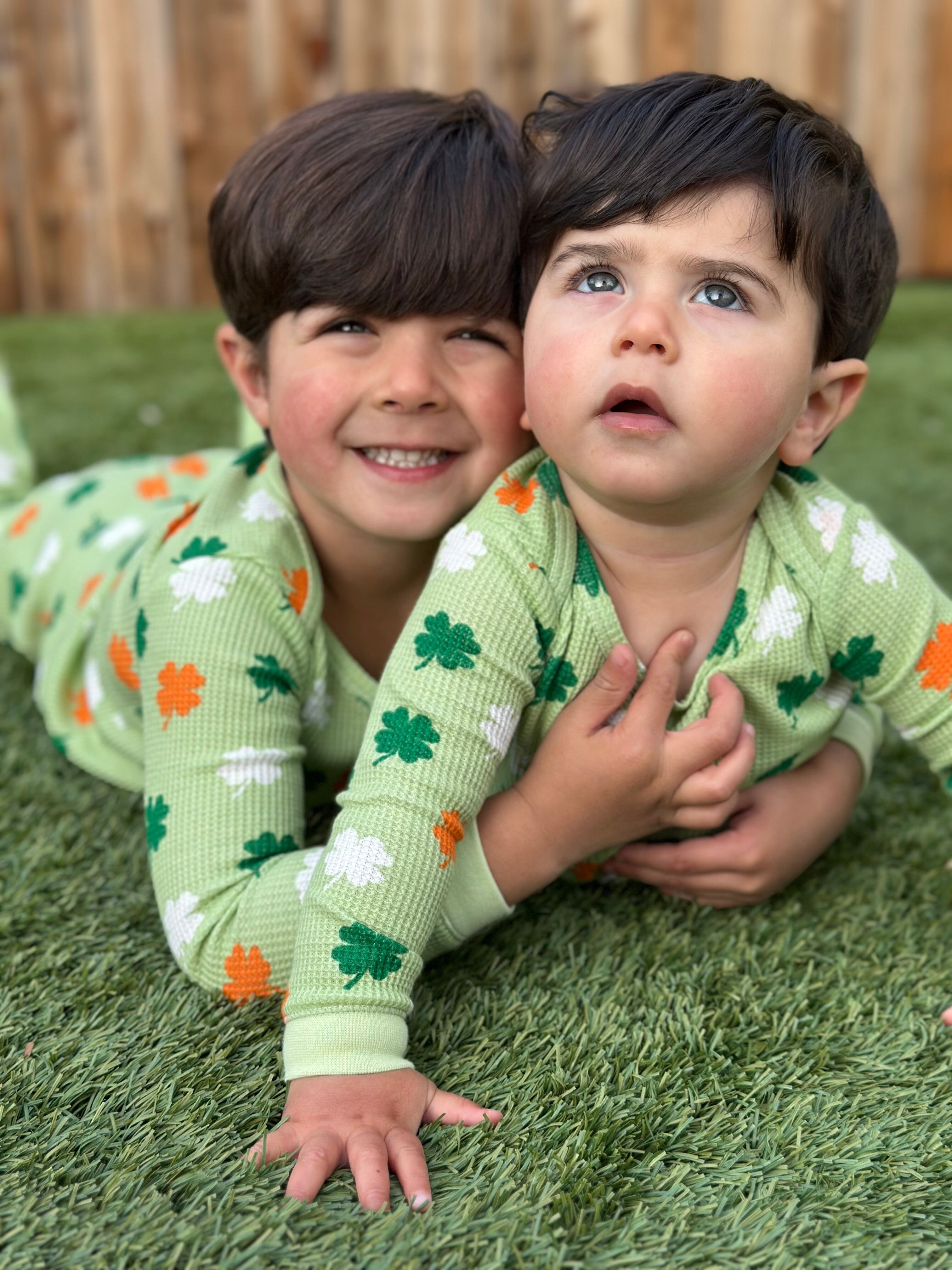 Two children in matching green pajamas with shamrocks lying on grass, smiling and looking upward.