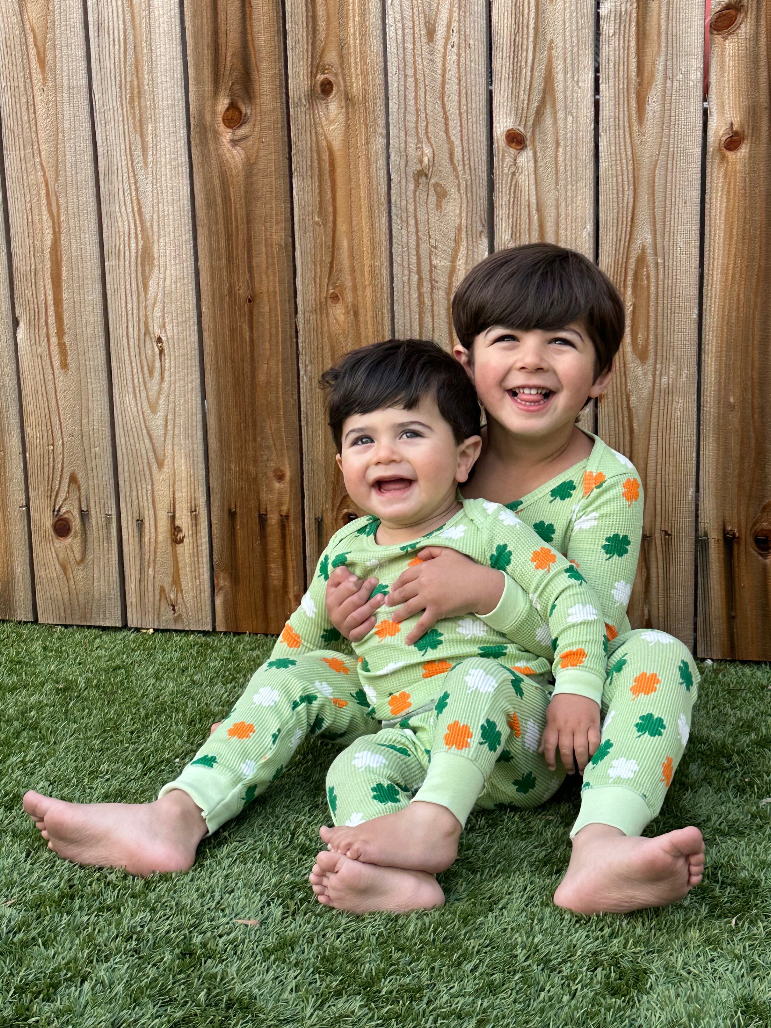 Two happy children in matching green pajamas with shamrock patterns sit on grass against a wooden fence.
