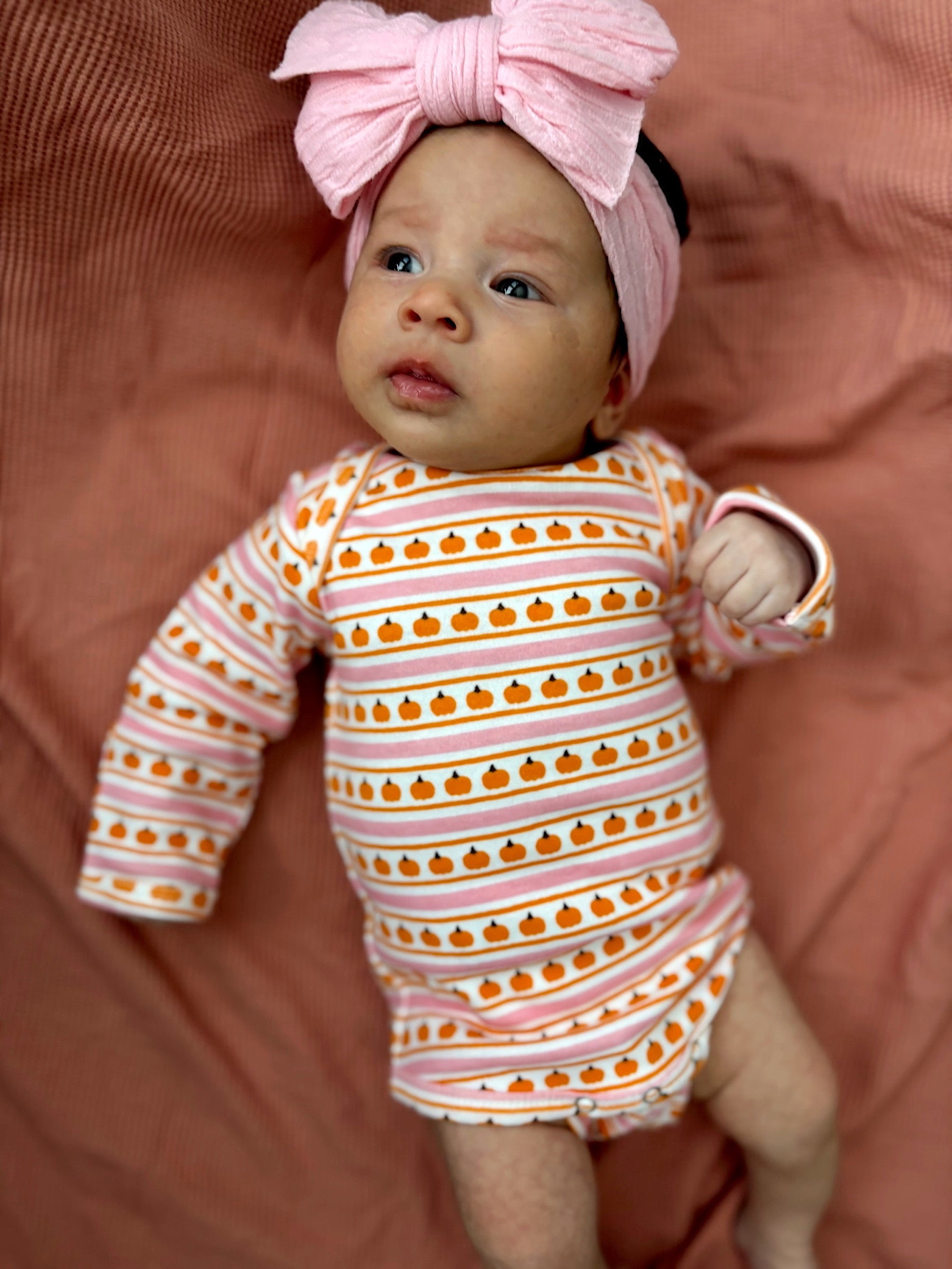 Baby in pumpkin-patterned onesie and large pink bow on a soft pink background.