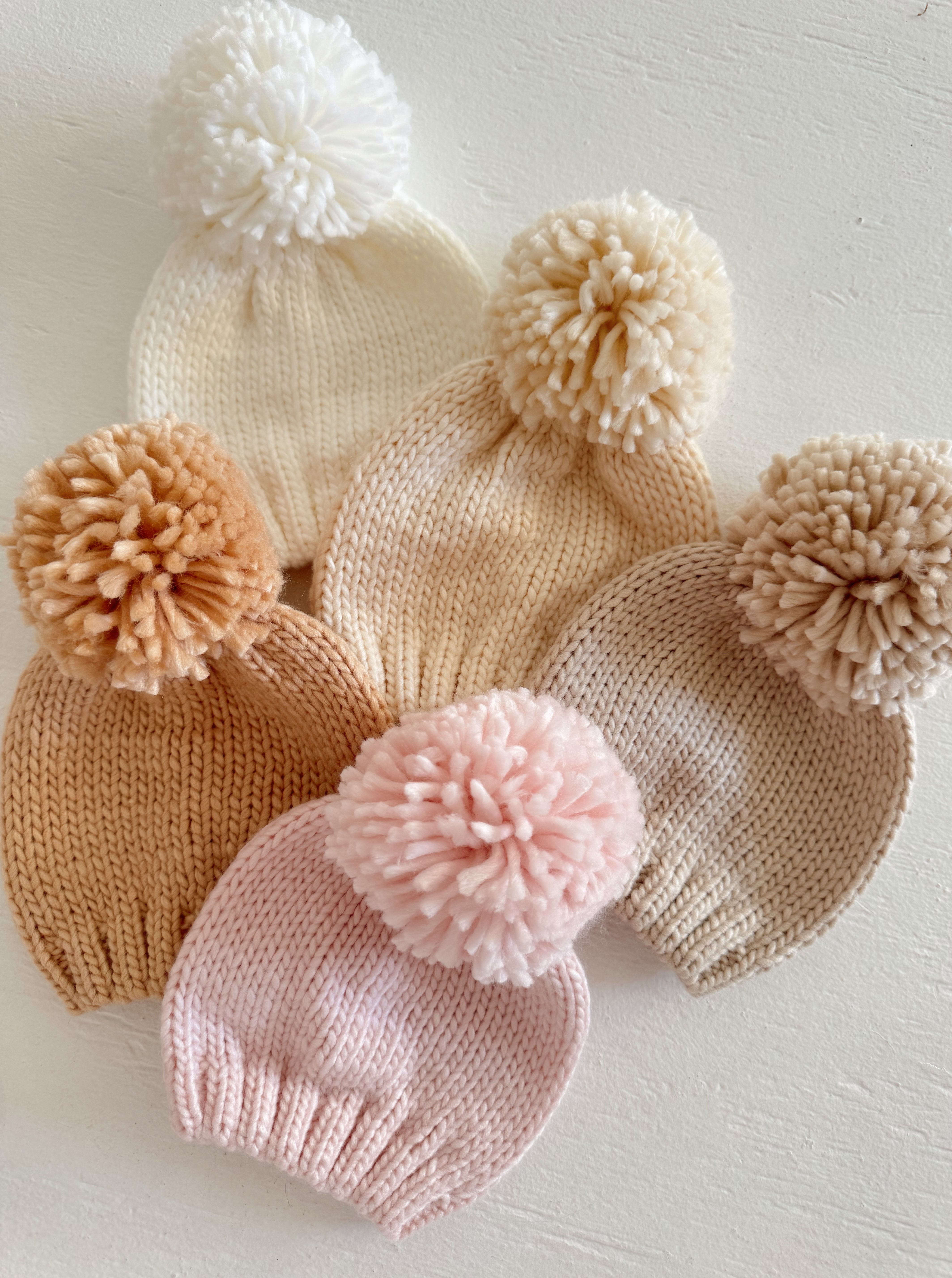 Assorted knitted beanies with pom-poms in cream, beige, and blush shades on a light background.