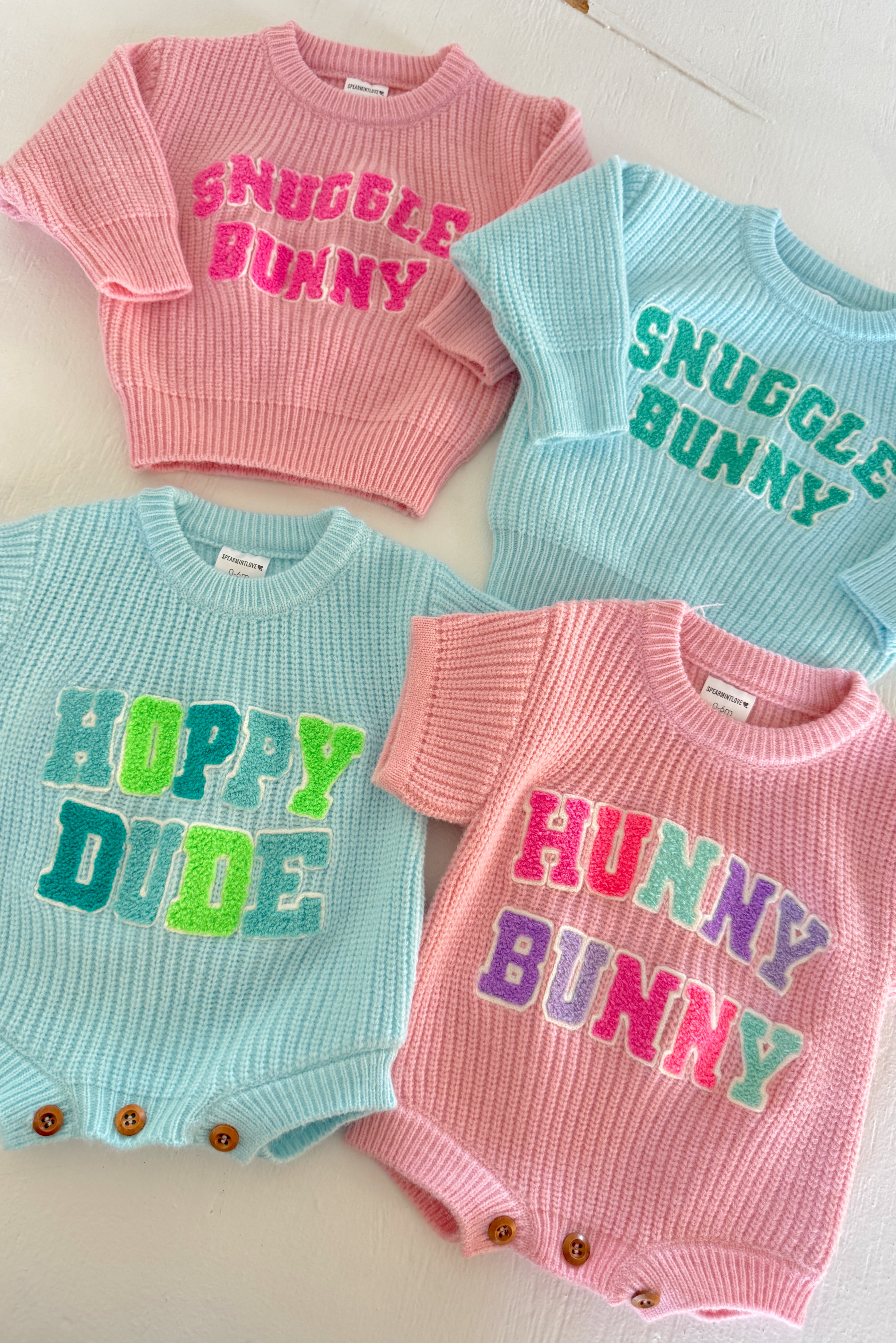 Four colorful infant sweaters displayed: pink with "Snuggle Bunny," blue with "Hoppy Dude," and more, on a white background.