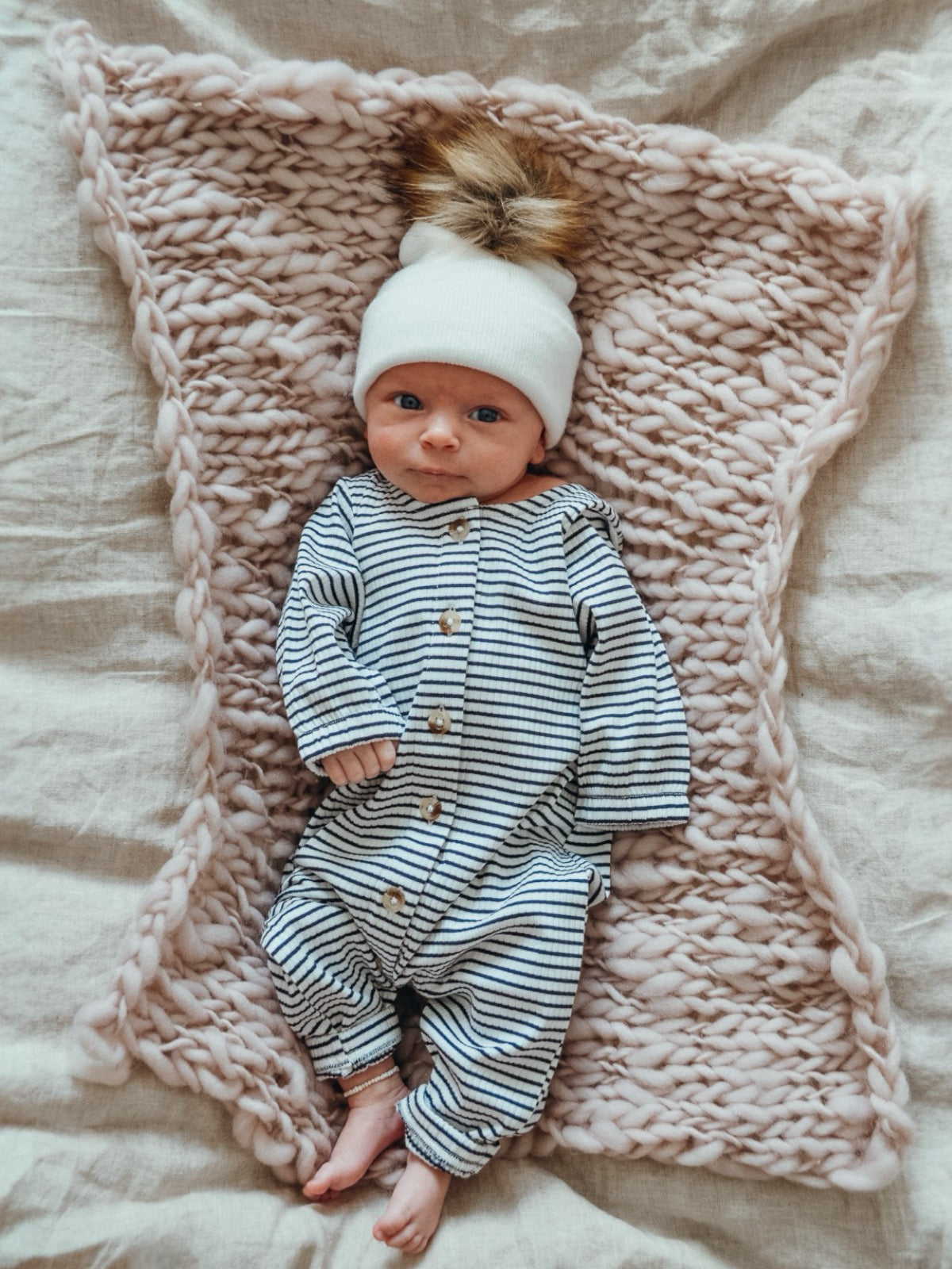 Infant in striped onesie and white hat, lying on a pink knitted blanket.