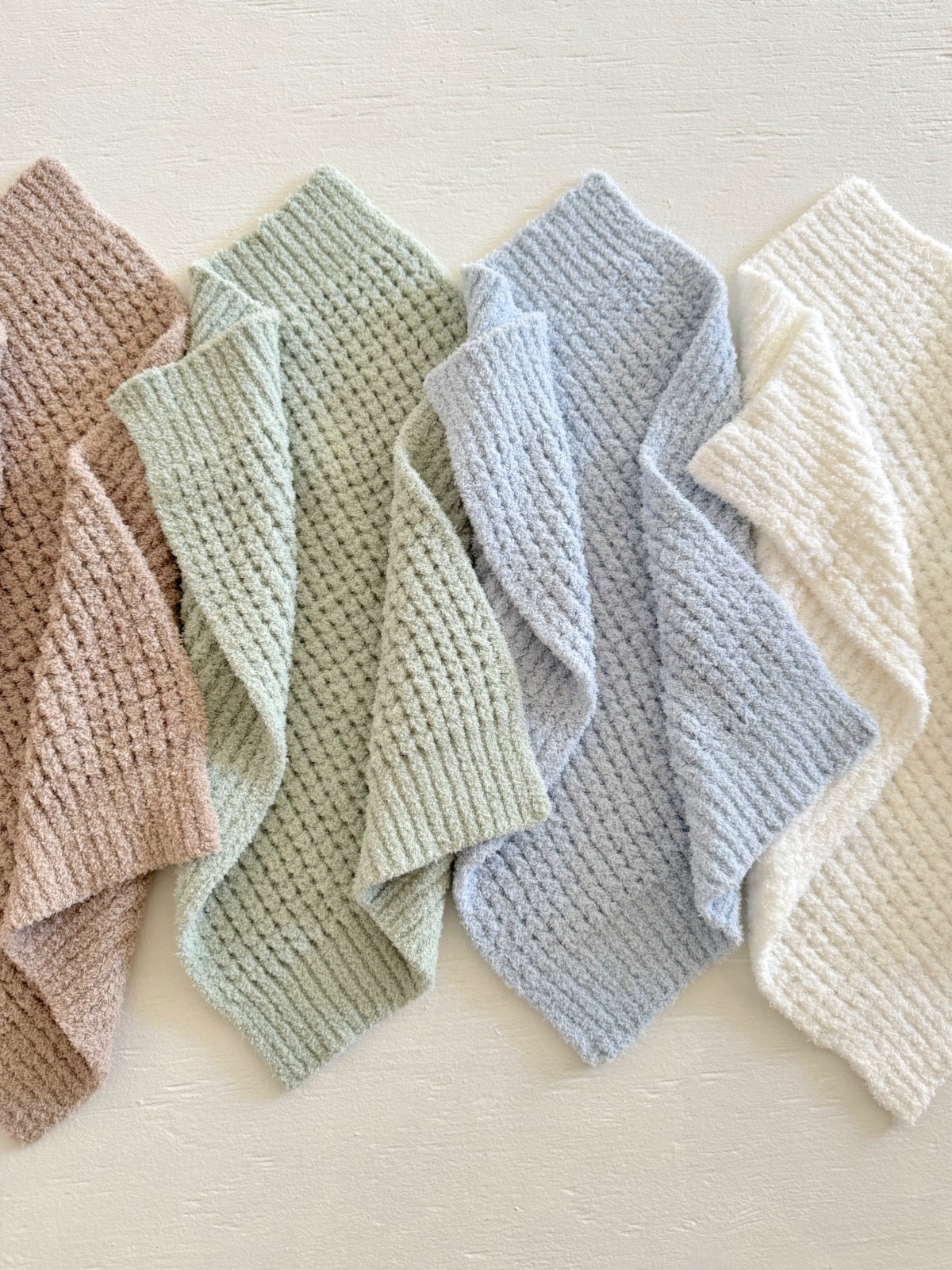 Four textured blankets in varying pastel colors: blush, green, blue, and cream, arranged neatly on a light background.