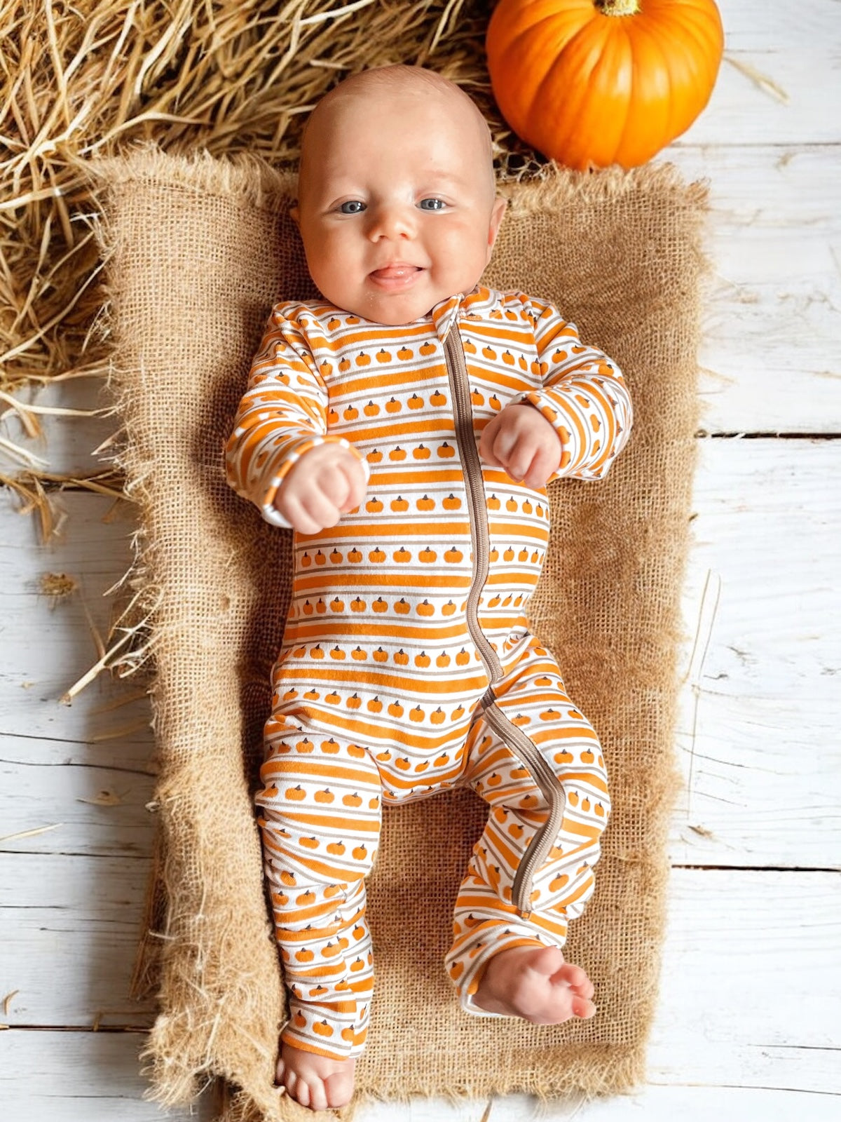 Smiling baby in pumpkin-patterned onesie on burlap with a pumpkin and hay background.