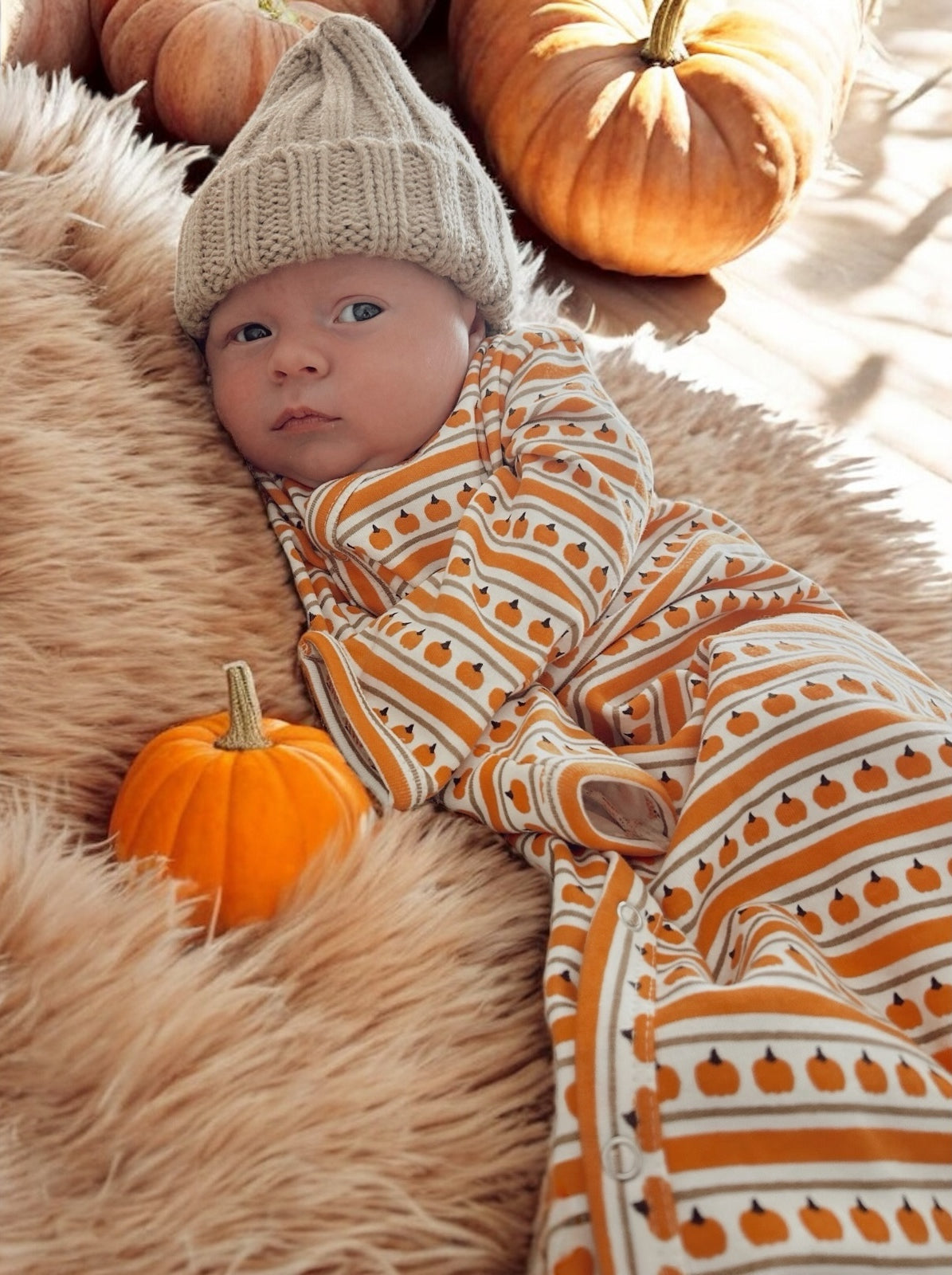 Baby in a pumpkin-patterned outfit, resting on fur with pumpkins in the background, exuding a cozy autumn vibe.