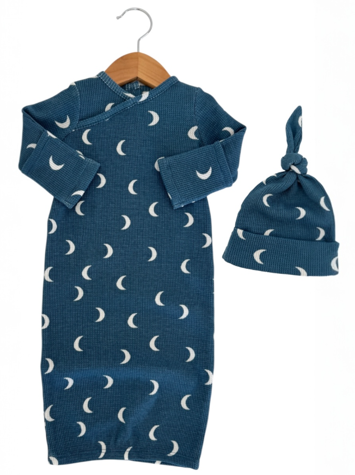 Blue baby gown with moon patterns and matching knotted hat, hung on a wooden hanger. Ideal for newborns.