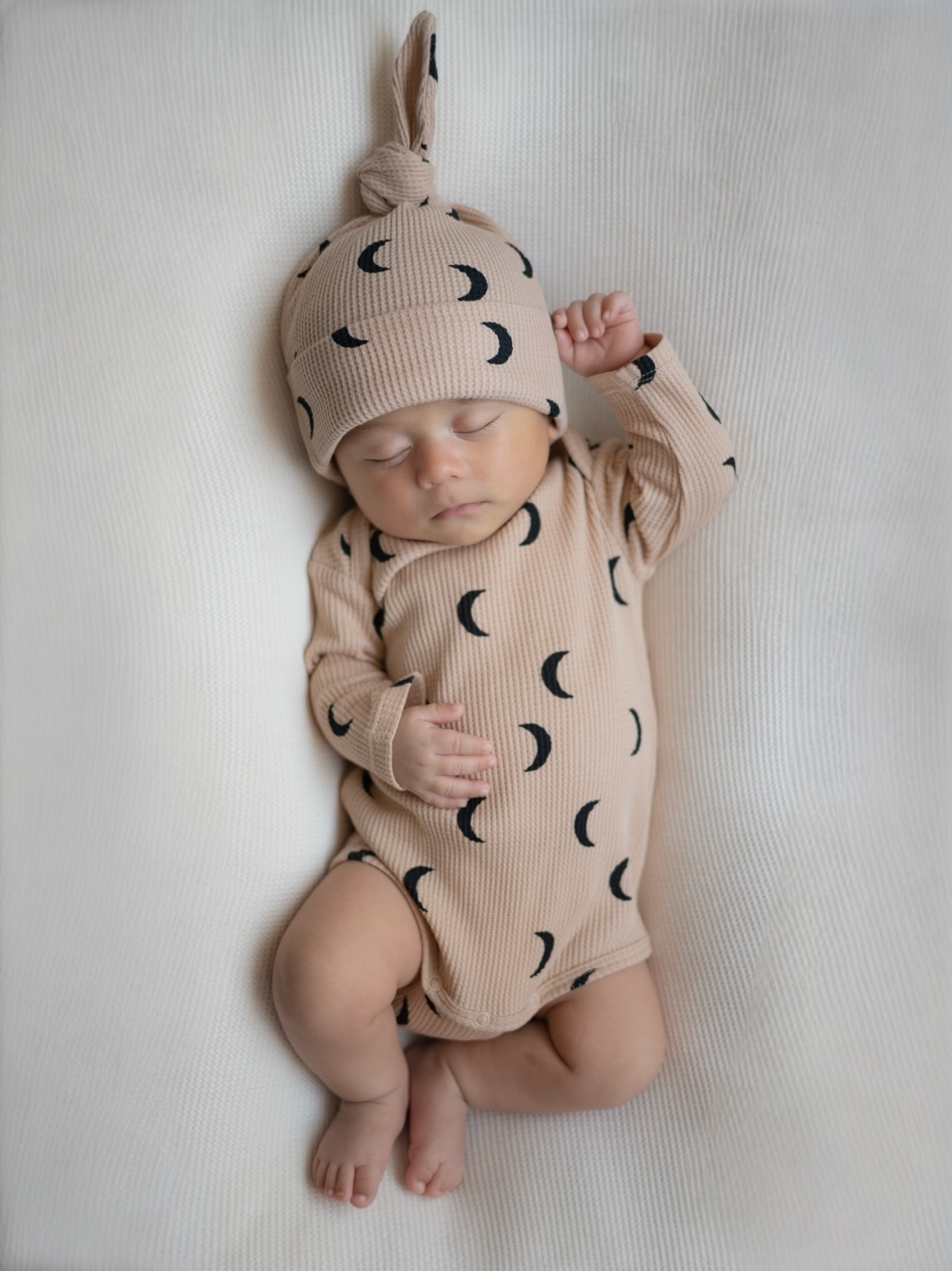 Sleeping baby in a crescent moon patterned onesie and hat on a textured white blanket.