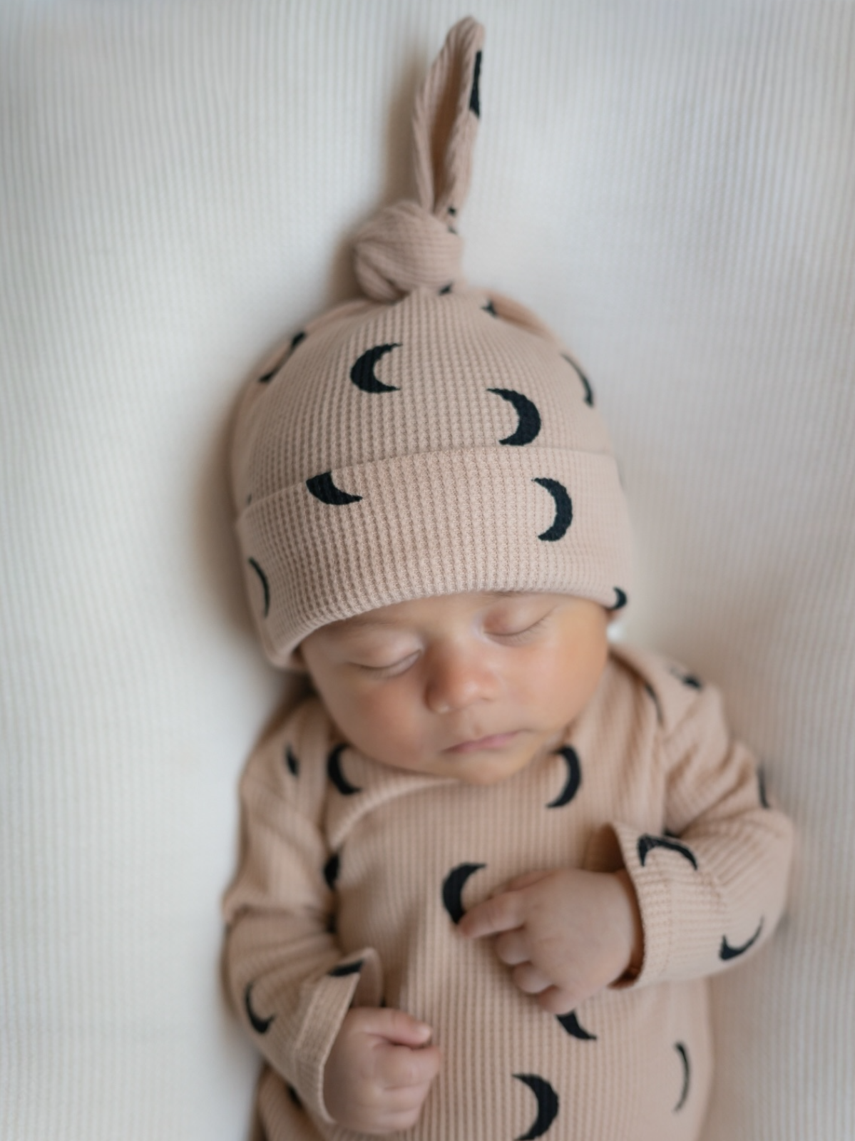 Sleeping baby wearing a soft, moon-patterned outfit and hat, lying on a textured white background.