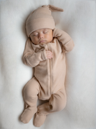 Sleeping baby in a tan outfit and matching hat, peacefully resting on a cream-colored blanket.