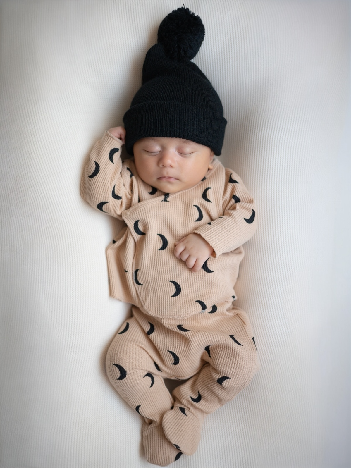 Sleeping baby in cozy moon-patterned outfit and black beanie, nestled on a soft cream blanket.
