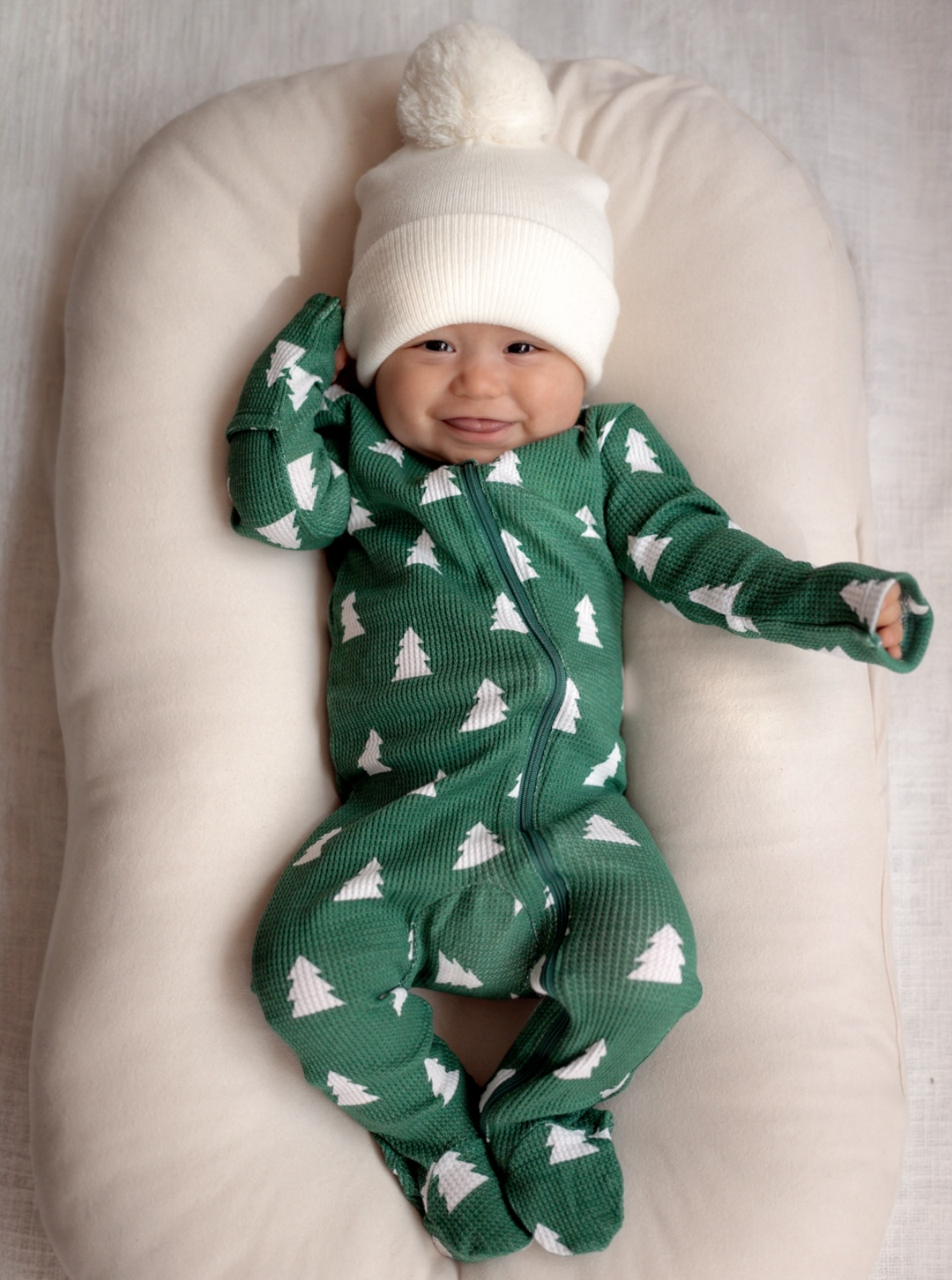 Smiling baby in a green onesie with trees, wearing a white pom-pom hat, resting on a soft pillow.