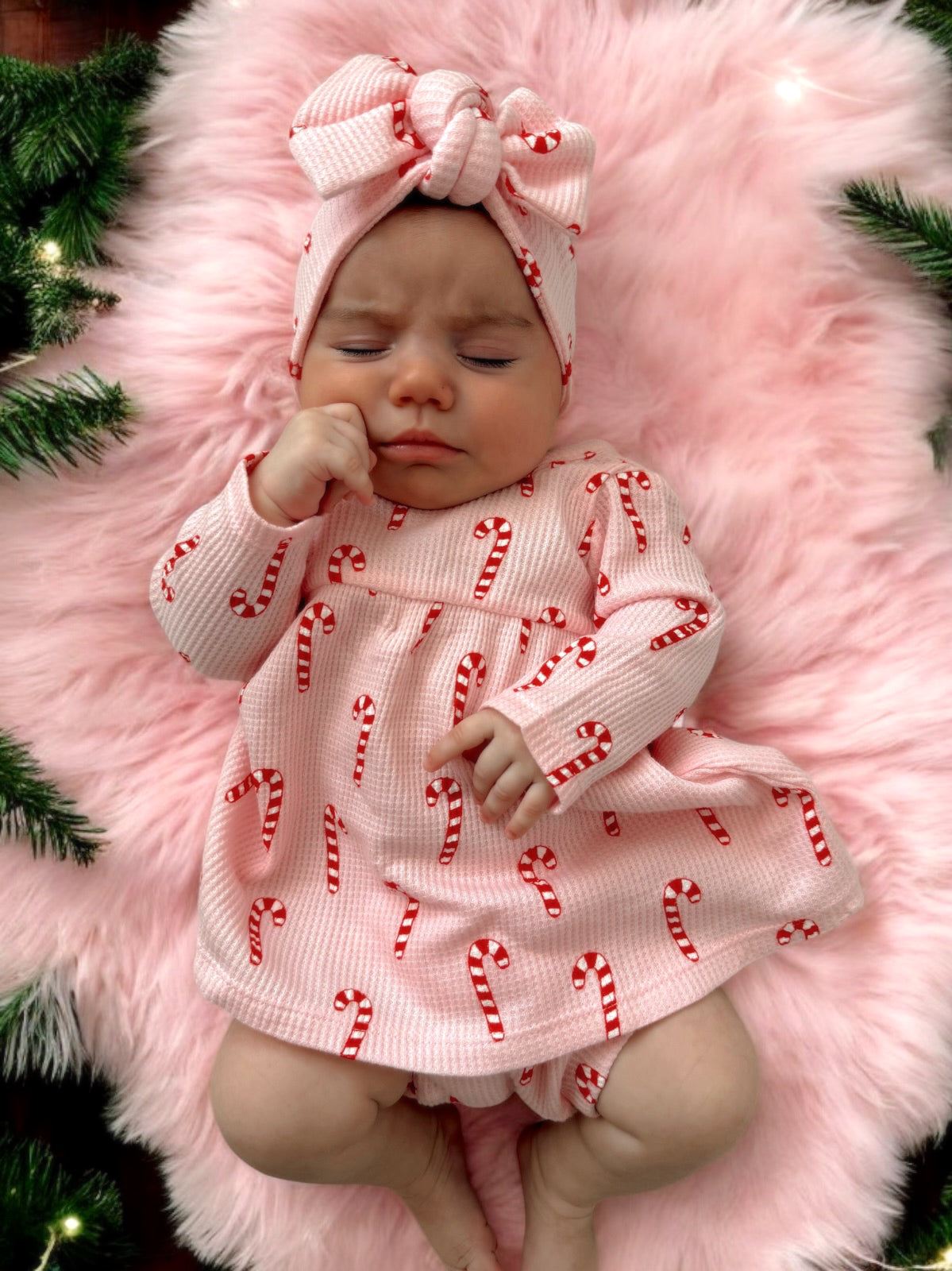 Baby girl in candy cane dress and headband, lying on pink fur with holiday greenery in the background.